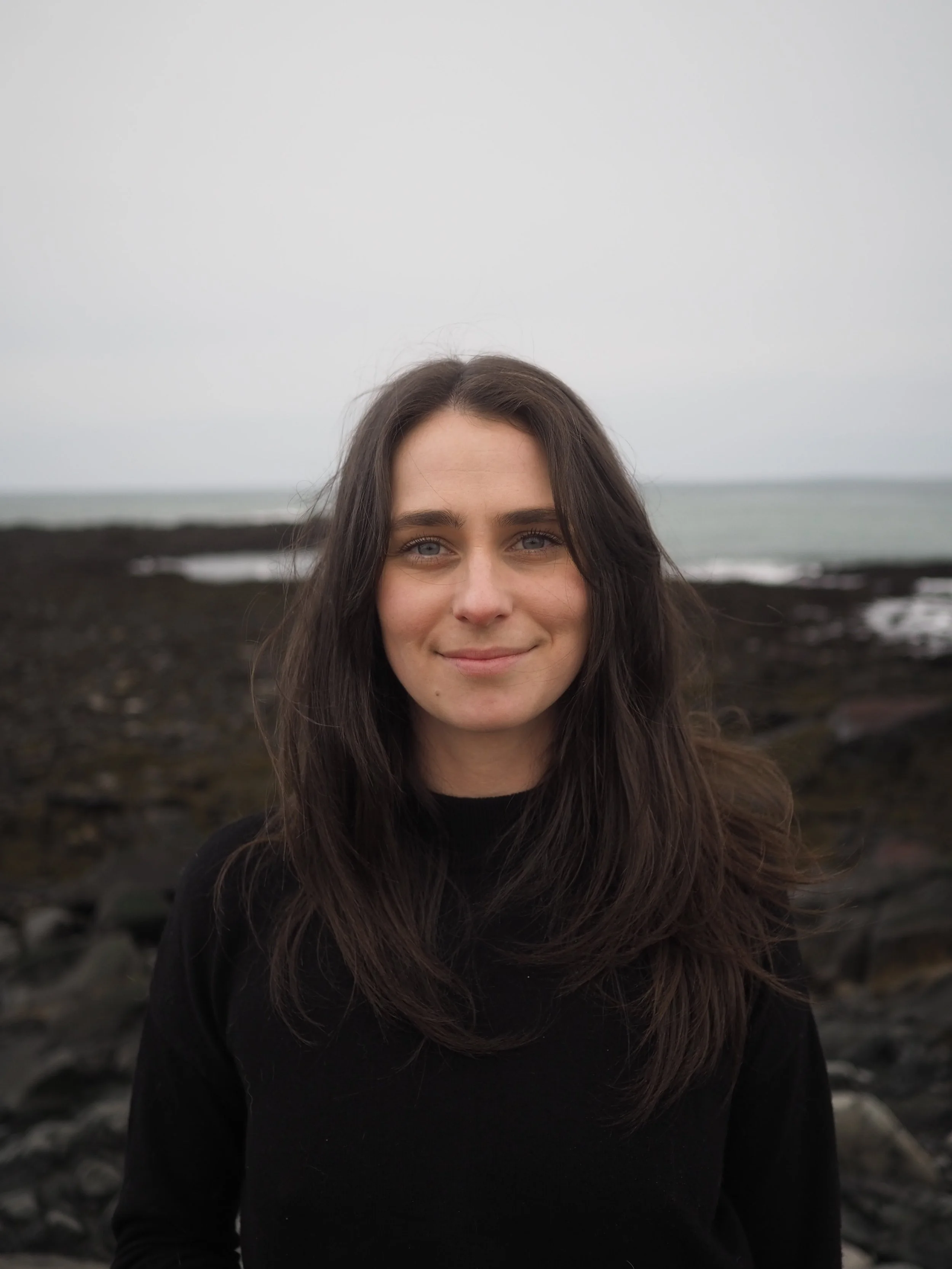 A woman with dark brown hair and blue eyes standing outdoors near a rocky shoreline on an overcast day.