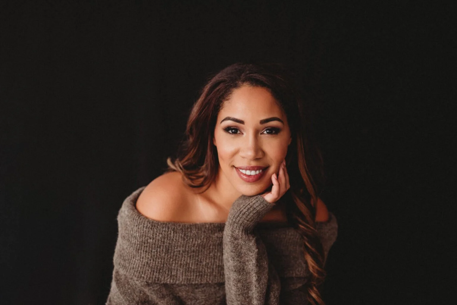 A young woman with long brown hair smiling, wearing a brown off-the-shoulder sweater, against a black background.