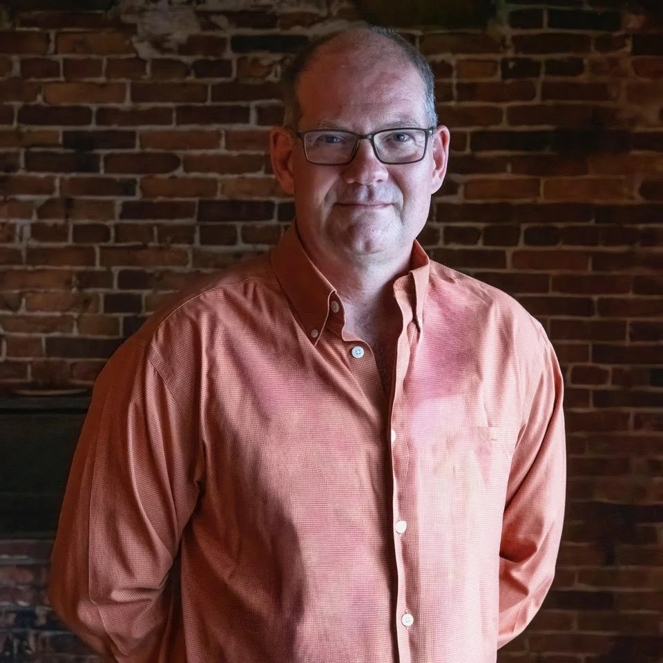 A middle-aged man with glasses and short hair, wearing an orange button-up shirt, standing in front of a brick wall.
