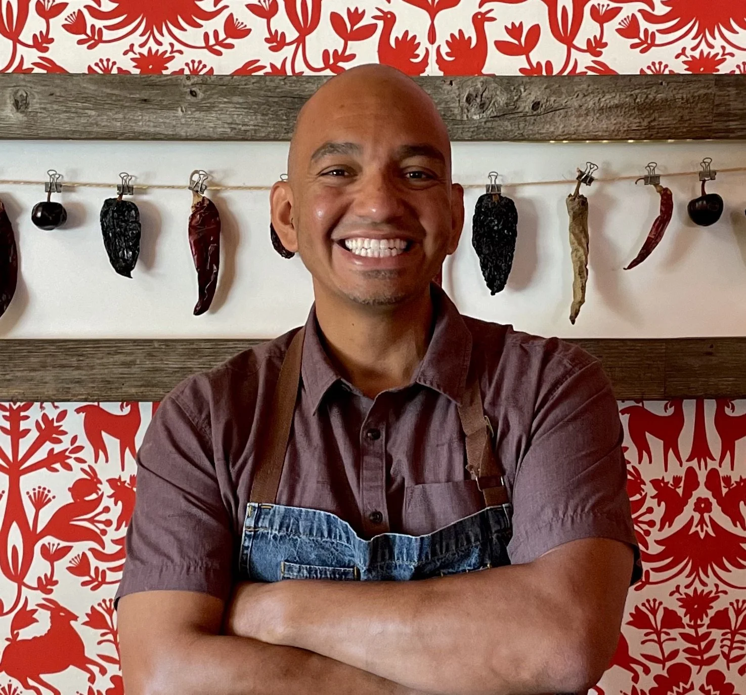 A smiling man with crossed arms wearing a brown shirt and apron, standing in front of a wall decorated with red and white floral and animal patterns, with dried peppers hanging on a string behind him.