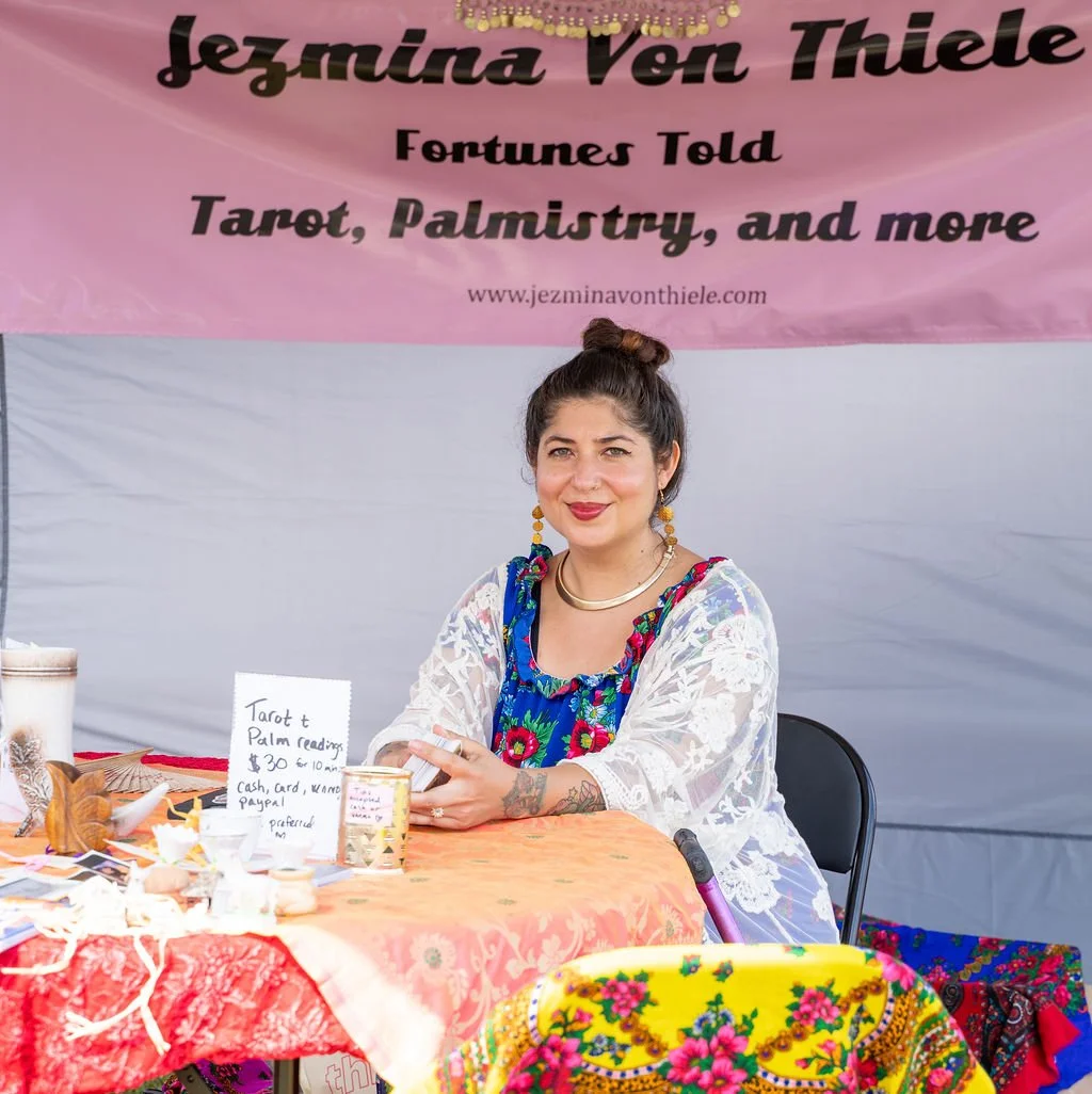 A woman with dark hair in a bun sitting at a booth with a pink banner that reads 'Jezmina Von Thiele, Fortunes Told, Tarot, Palmistry, and more'. She is smiling, wearing colorful earrings, a gold necklace, and a white lace shawl over a blue floral dress. The table in front of her has tarot cards, a sign offering tarot and palm reading services for $30 to $10, and various decorative items.