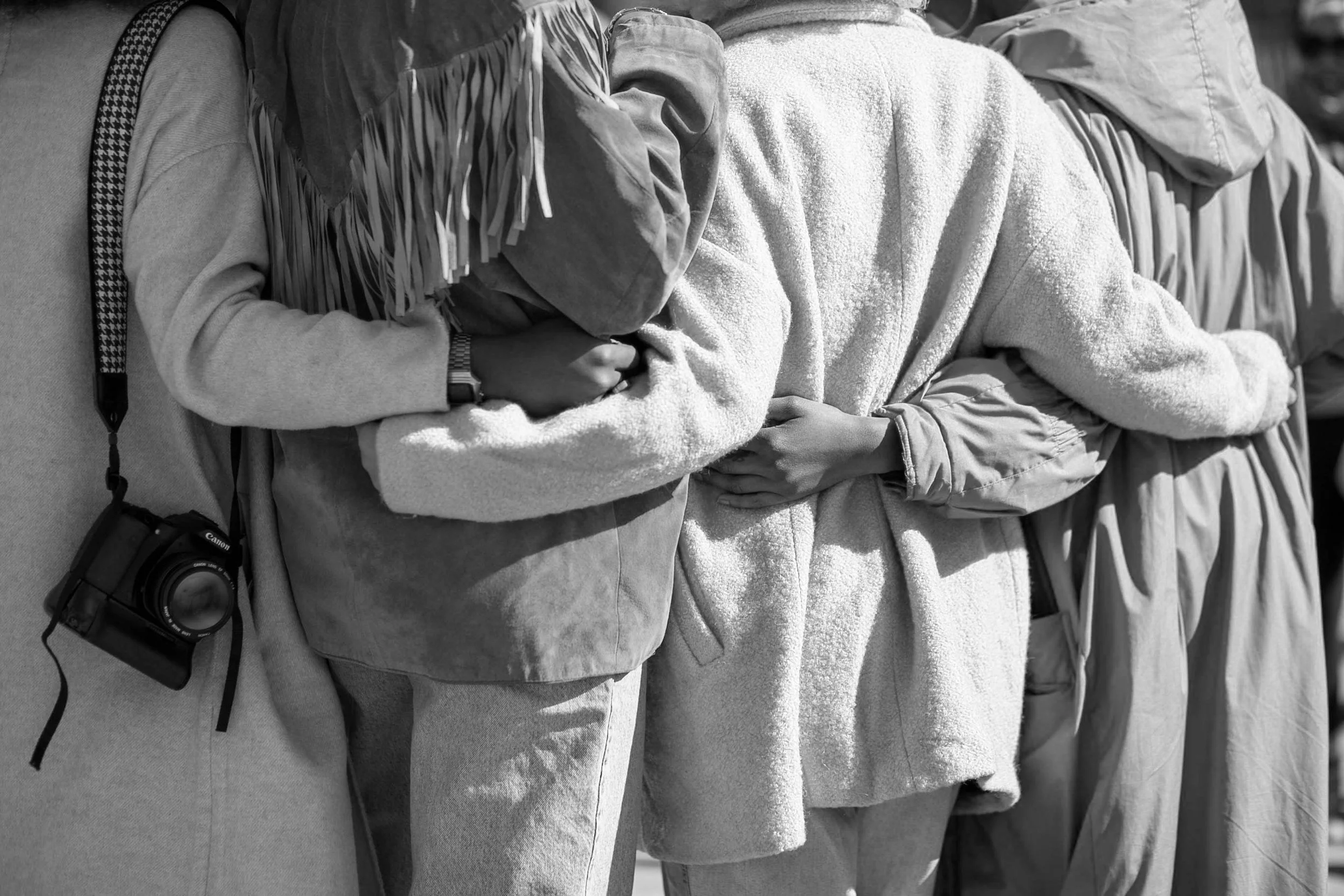 Three people standing in a line with their arms linked, holding their stomachs, wearing casual clothing in a black-and-white photo.