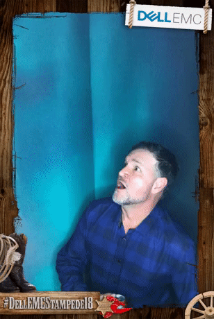 A man with dark hair and a beard wearing a blue plaid shirt standing in front of a blue backdrop. There is a sign that says 'Dell EMC' at the top right corner and a hashtag '#DellEMCStampede18' at the bottom left corner.