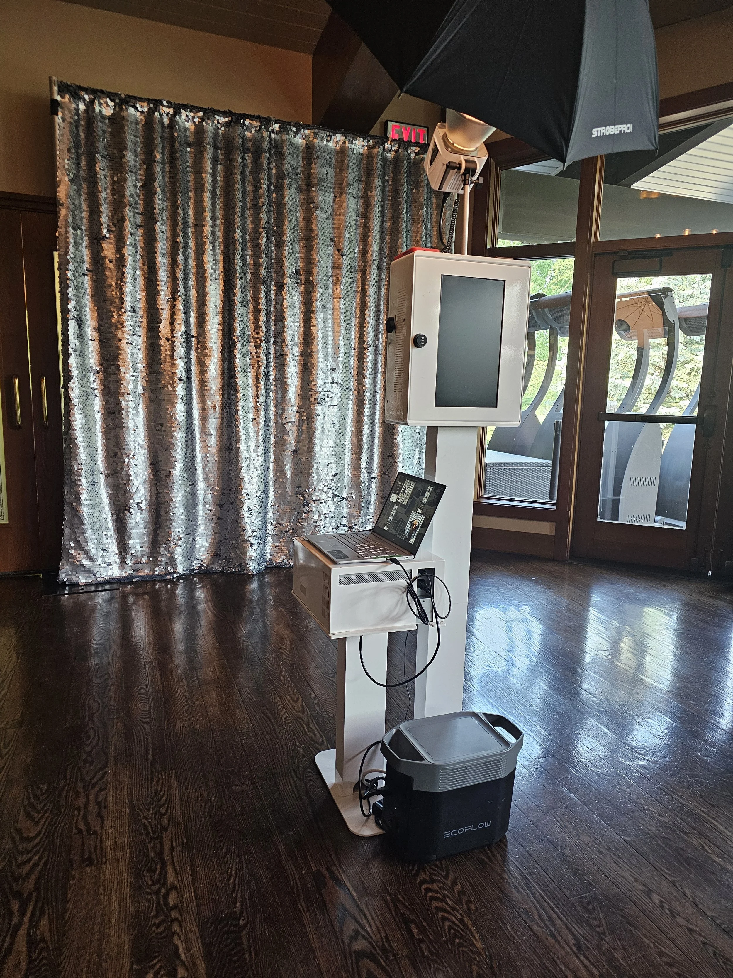 Photo of a photo booth setup inside a room with wooden floors, a silver sequin curtain backdrop, a large black umbrella, a monitor, a laptop, and a cooler.