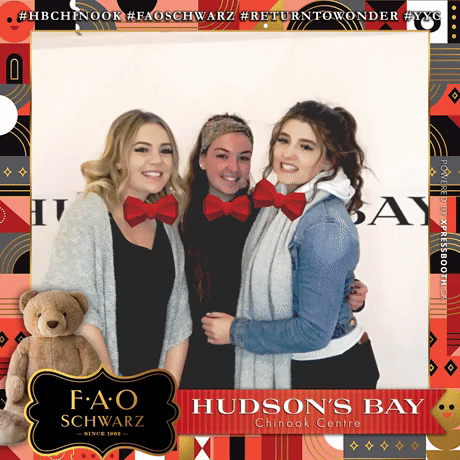 Three women wearing red bow ties smiling and standing close together at a Hudson's Bay event inside the Chinook Centre, with a teddy bear and decorative border in the photo.