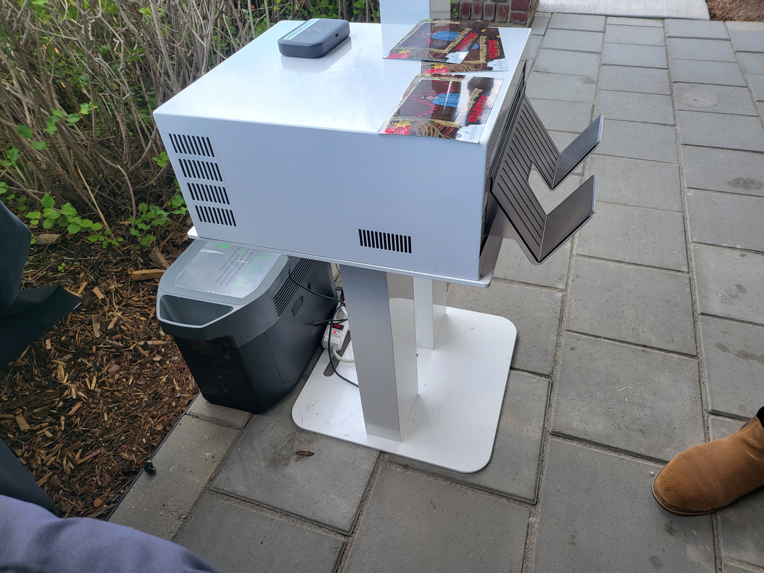 A white table with a small black computer tower underneath, placed on a concrete sidewalk outside. On top of the table are magazines and a small rectangular device, with two metal chairs attached to the side of the table. To the left, there's a bush