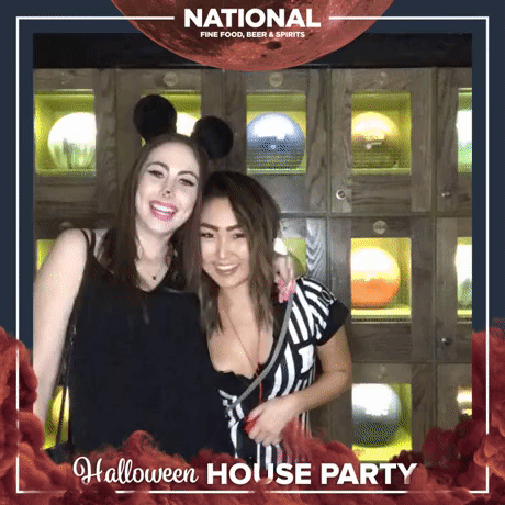 Two women at a Halloween house party, smiling and posing for a photo, with a wooden wall featuring illuminated globe decorations in the background.
