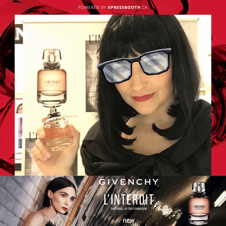 Woman with black hair and glasses holding a bottle of perfume at a promotional display for Givenchy L'Interdit perfume.
