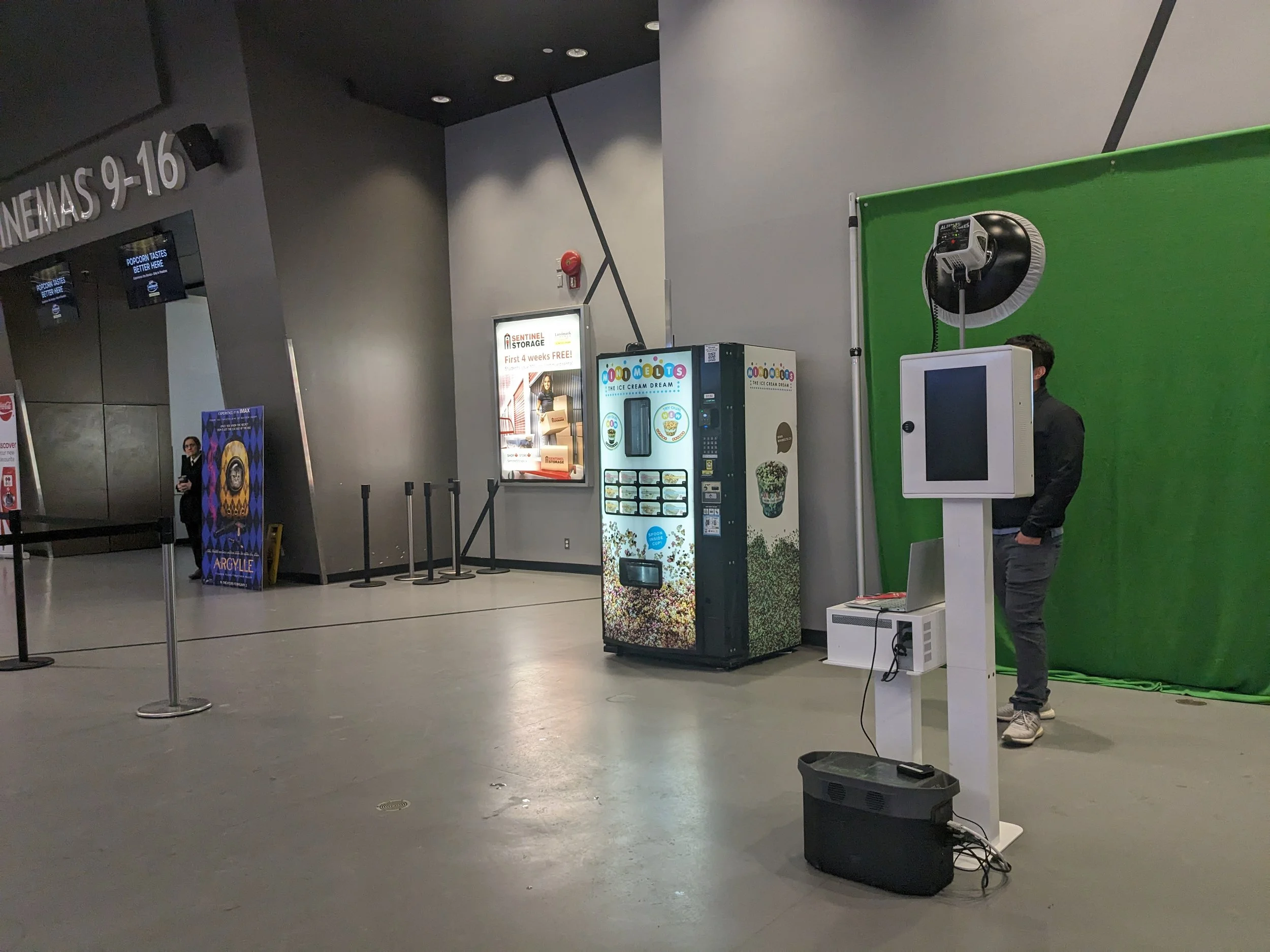 A person standing in front of a green screen setup in a lobby area with vending machines and a movie theater entrance in the background.
