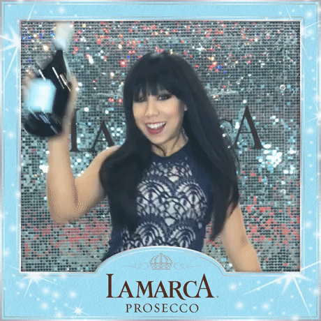 Woman with long black hair and bangs in a lace navy dress smiling and holding a bottle of champagne in front of a sparkly background, inside a blue decorative frame with 'Lamarca Prosecco' logo.