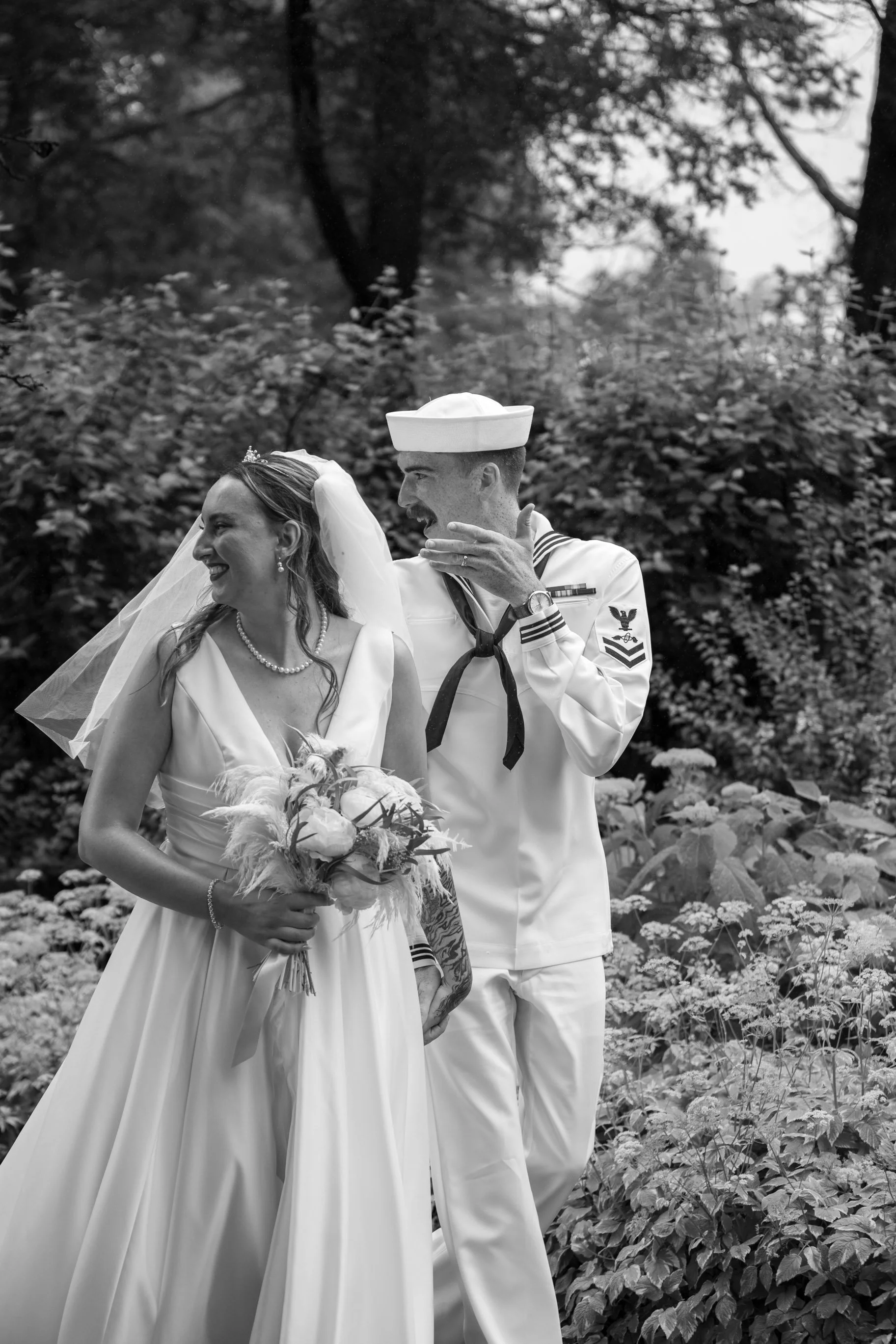 A bride in a wedding dress and a groom dressed as a sailor in a naval uniform, smiling and looking at each other, standing outdoors in front of shrubbery and trees.
