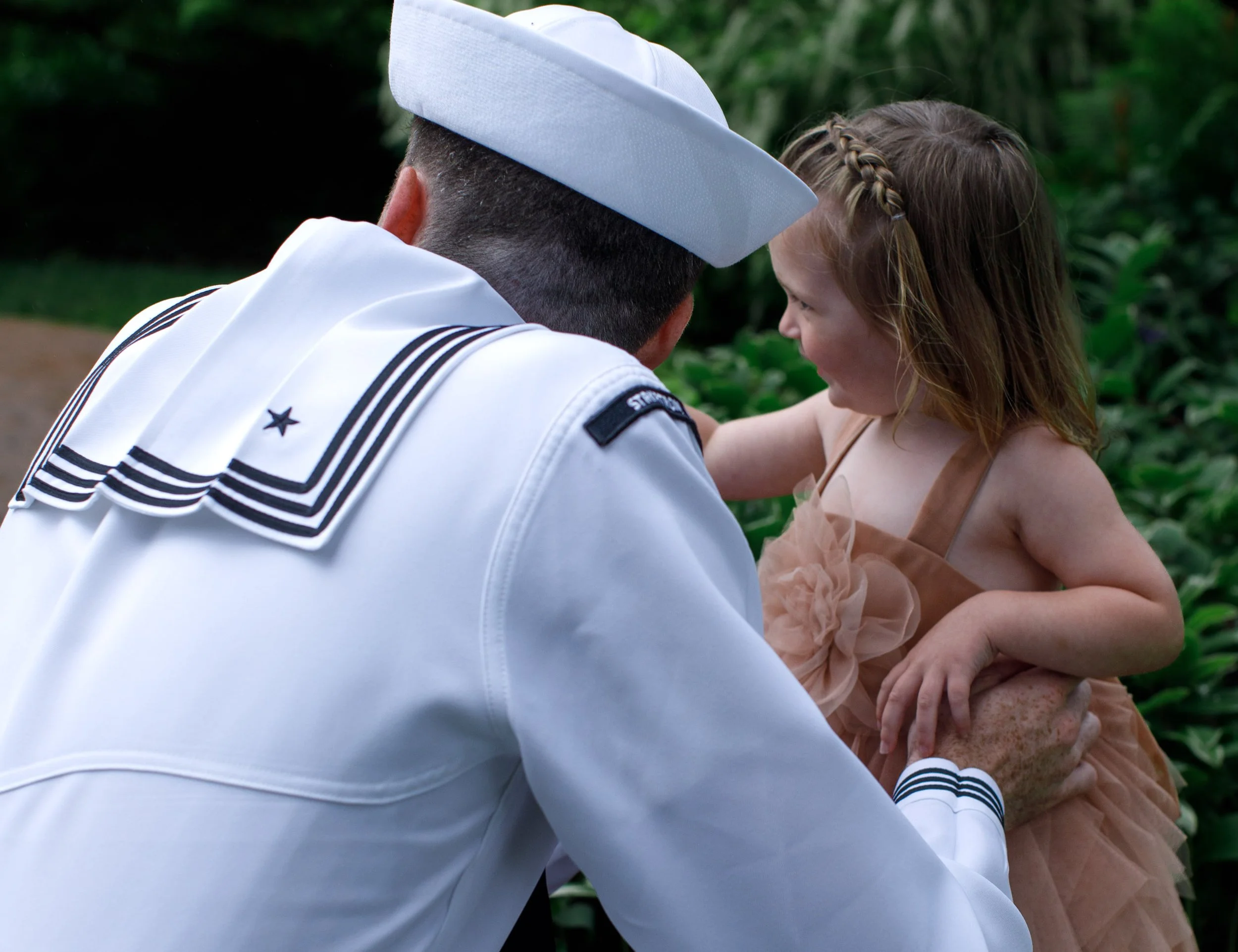 A sailor in a white uniform and hat holding a young girl in a dress, outdoors with green foliage in the background.