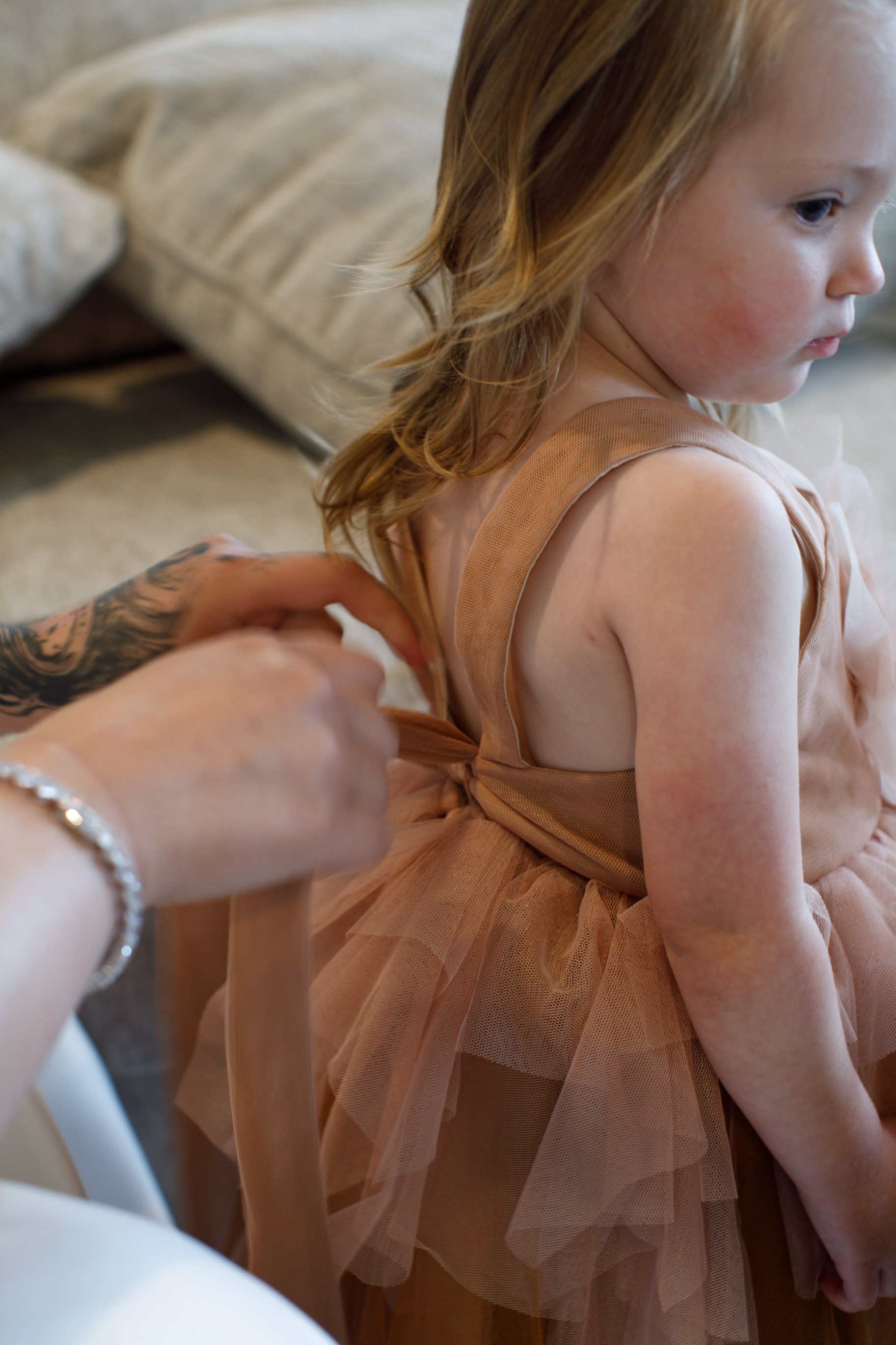 A young girl with brown hair is getting dressed in a peach-colored tulle dress while an adult helps tie the back.