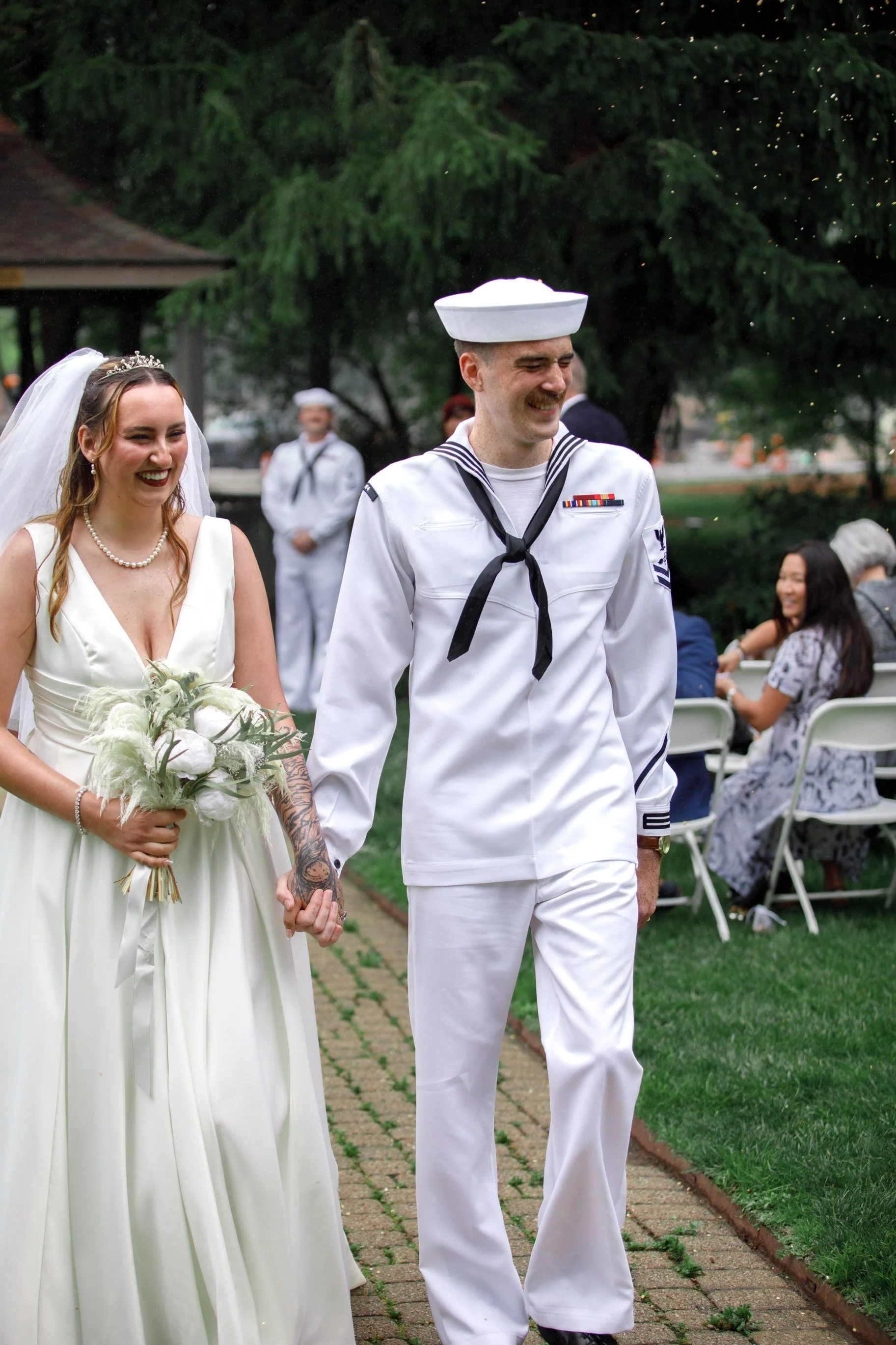 A couple, with the bride in a white wedding dress and the groom in a U.S. Navy sailor uniform, holding hands and walking outdoors while smiling, with guests seated at tables in the background.