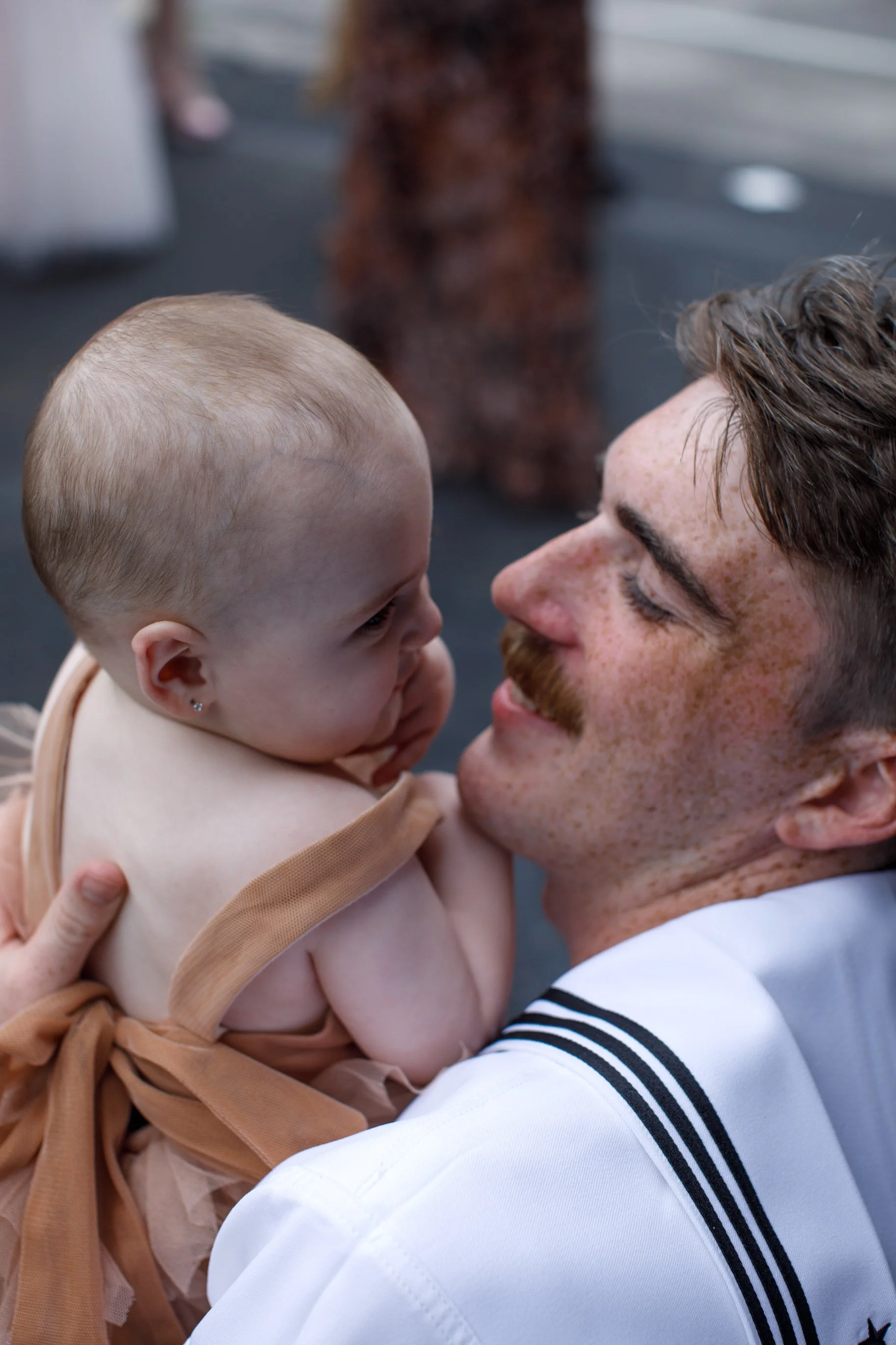 A man with a mustache and freckles holds a young girl with a shaved head close, both smiling and touching foreheads in an affectionate moment.