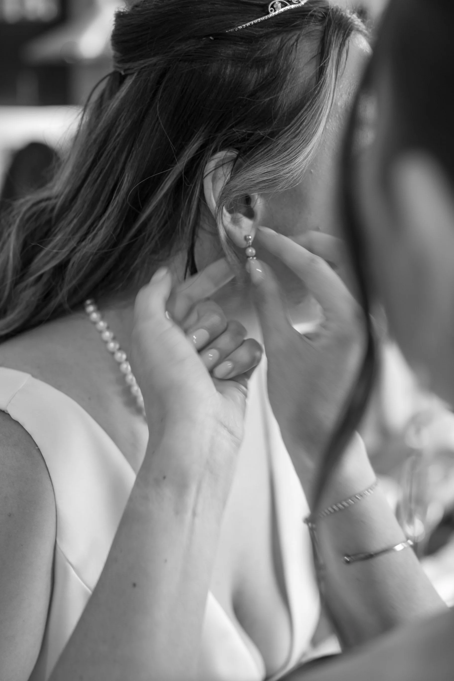 A woman in formal attire adjusting her earring, with jewelry including a pearl necklace, earrings, bracelet, and ring, captured in black-and-white.