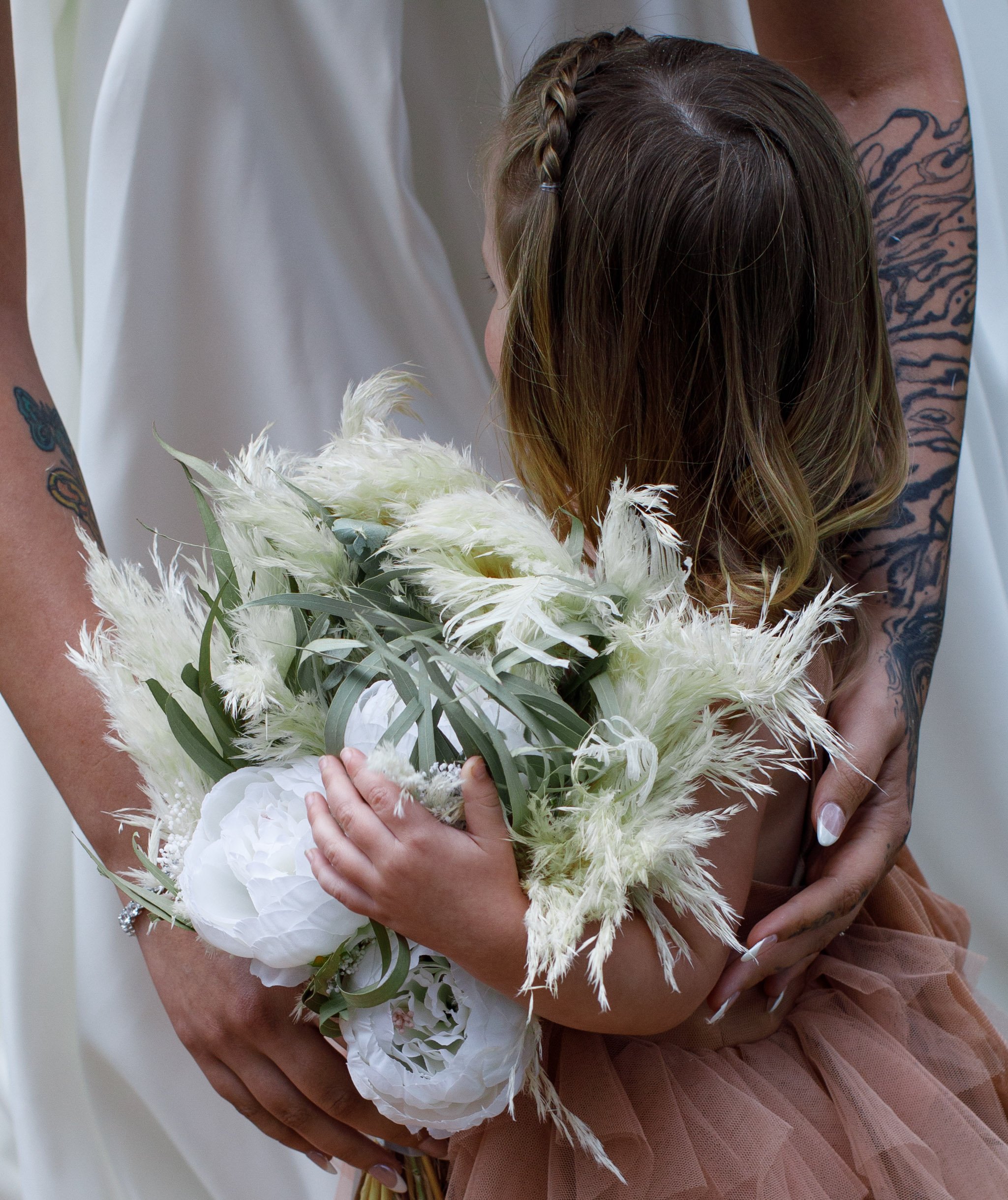A young girl with long brown hair, holding a bouquet of white flowers, being embraced by an adult, likely during a wedding or special event.