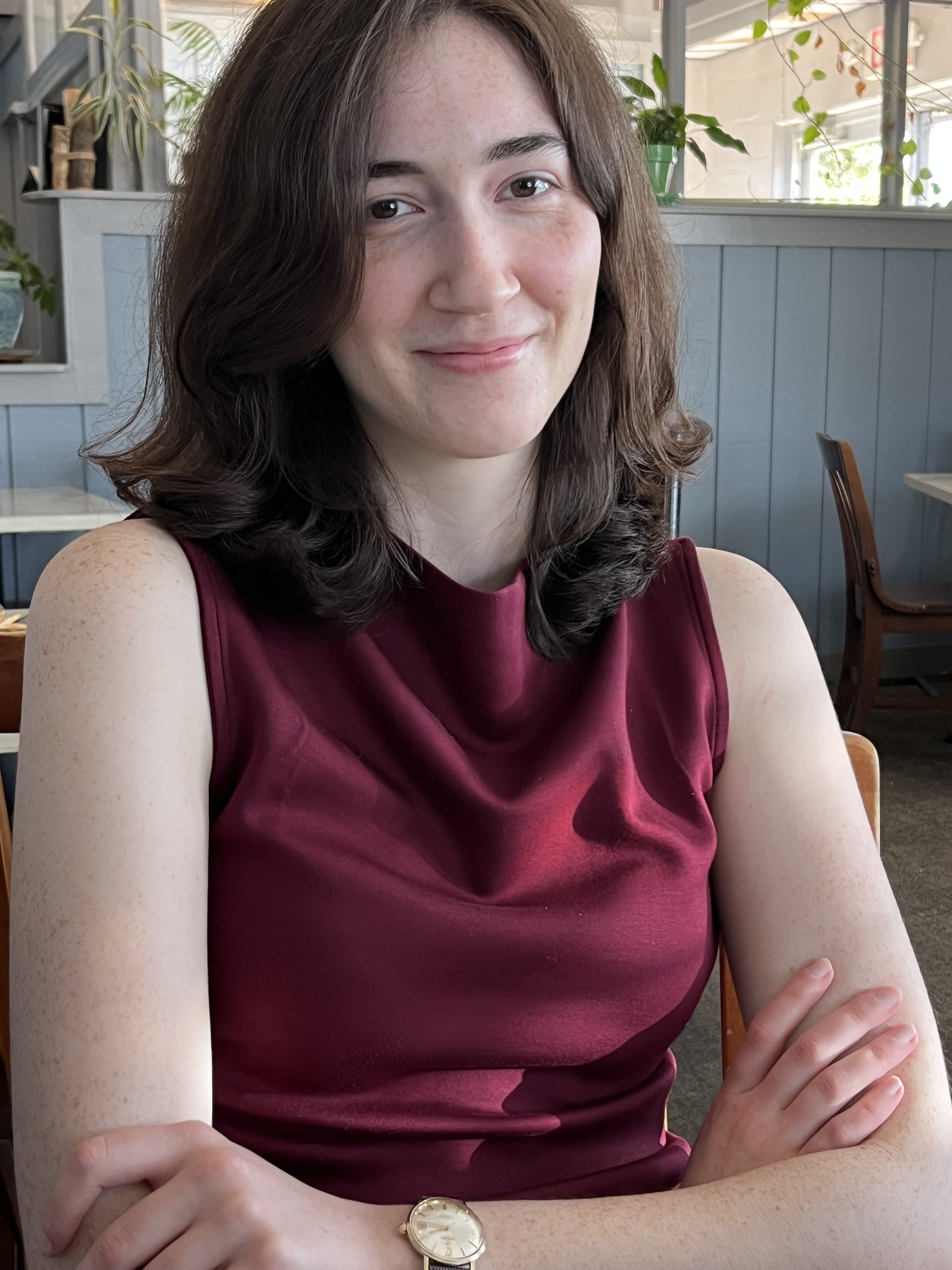A woman with shoulder-length brown hair, wearing a sleeveless maroon top, sitting at a table inside a restaurant with a blue wall and plants in the background.