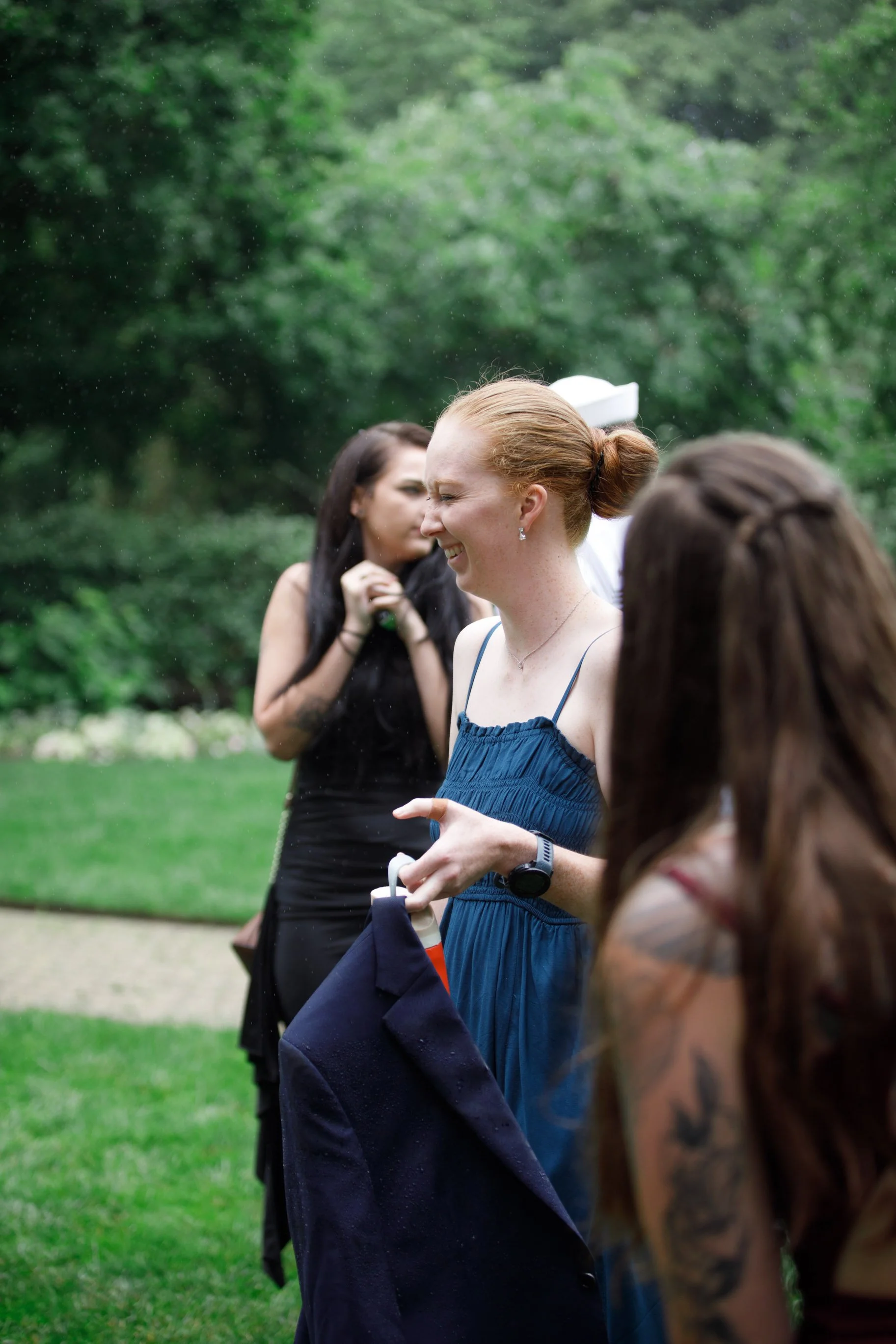 Three women are outdoors on a grassy area with trees, smiling and laughing while holding drinks, in a casual gathering during light rain.