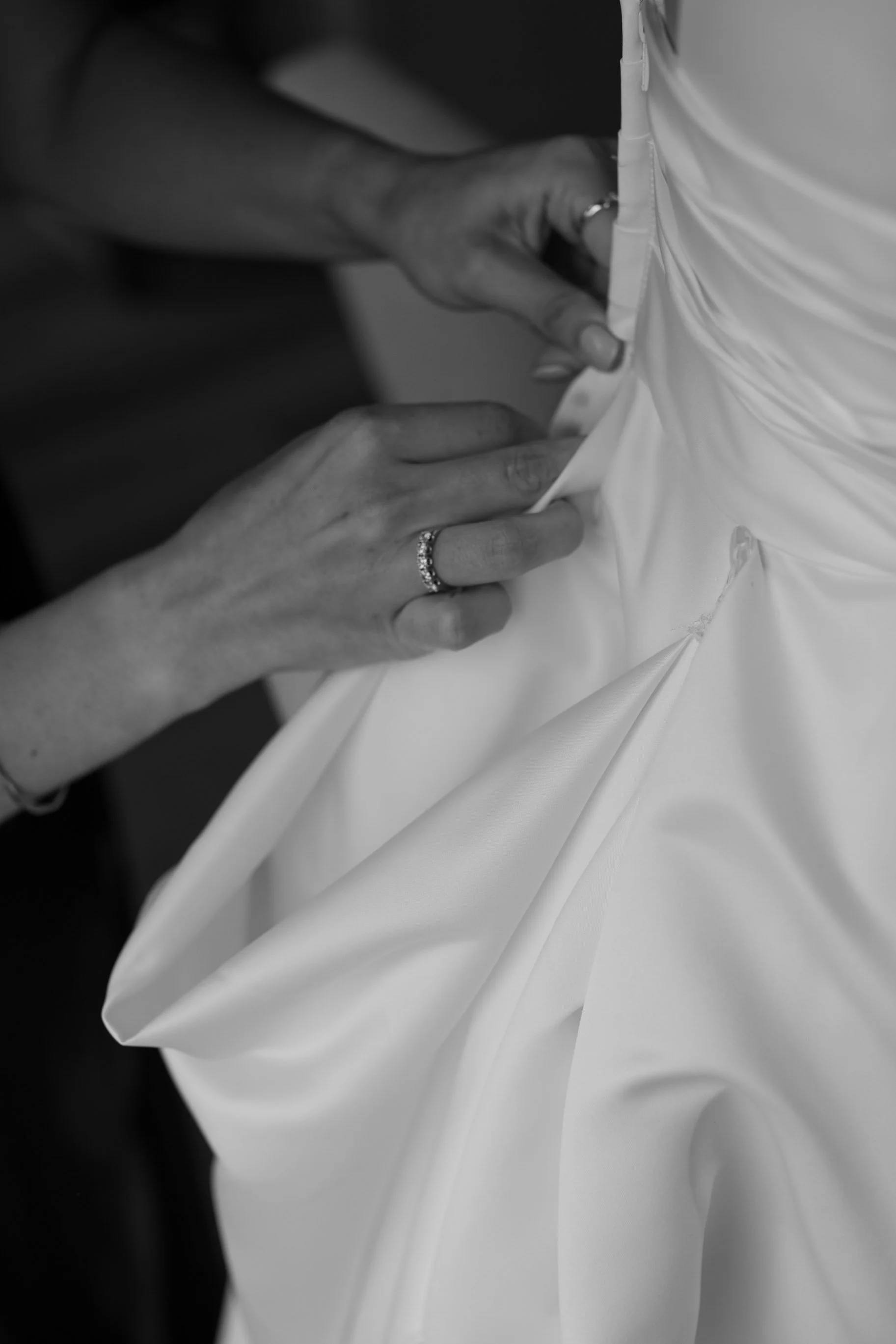 Close-up black-and-white photo of a woman adjusting a wedding dress, showing her hands with rings on her fingers.