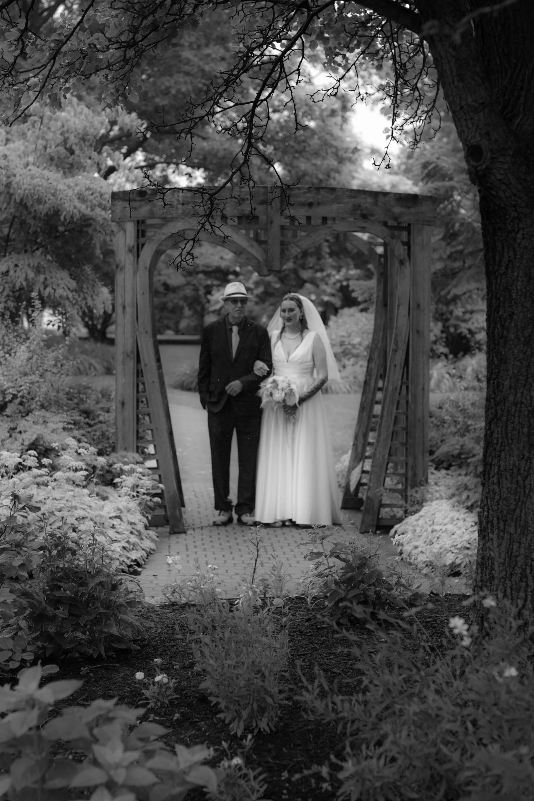 Black-and-white photo of a bride and an older man walking together under a wooden heart-shaped archway in a garden. The bride is in a long white wedding dress holding a bouquet, and the man is in a suit and hat.
