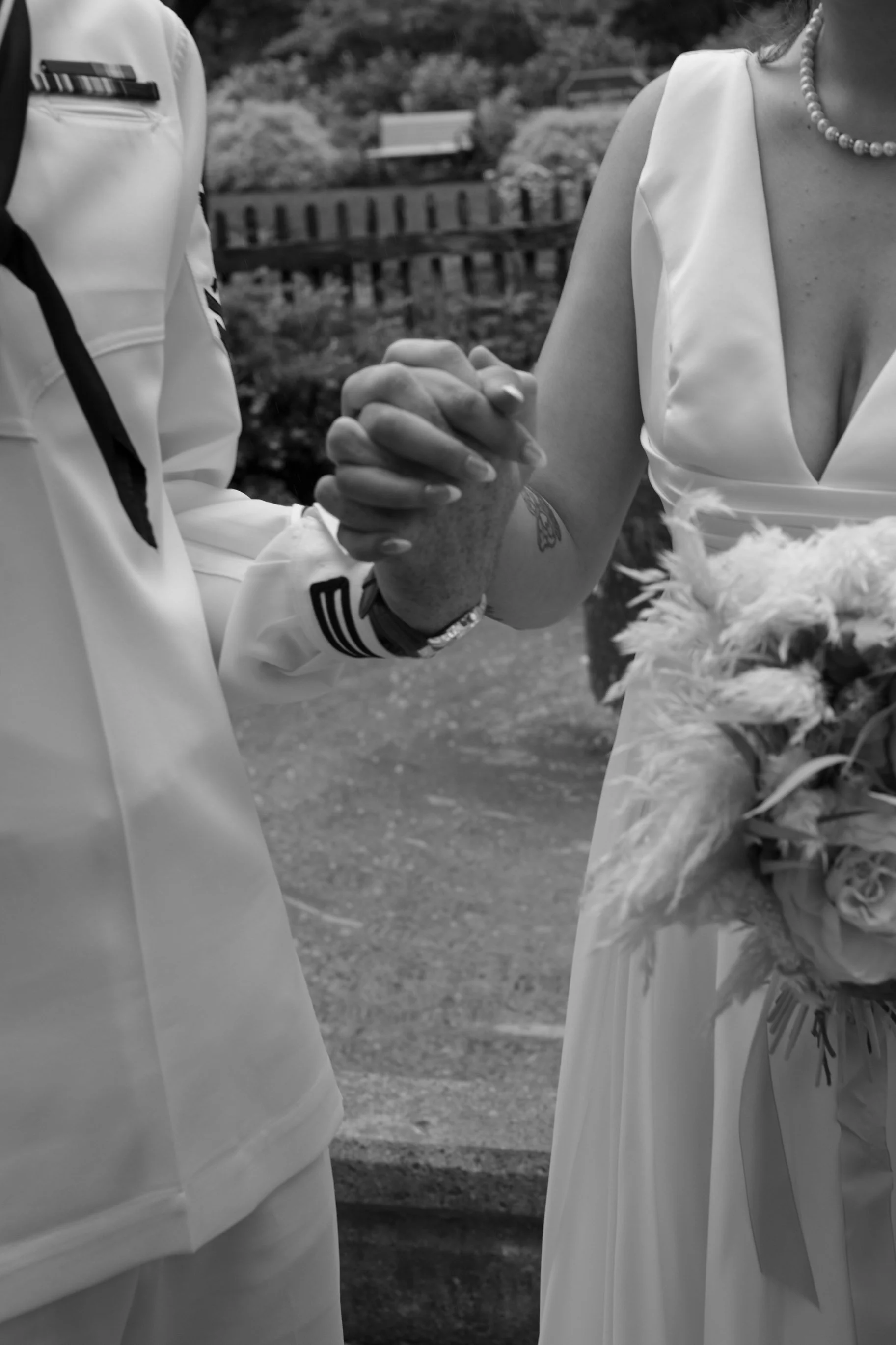 Close-up of a wedding couple holding hands, with the groom's hand gently clasped with the bride's. The bride is holding a bouquet and wearing a sleeveless dress with a pearl necklace.