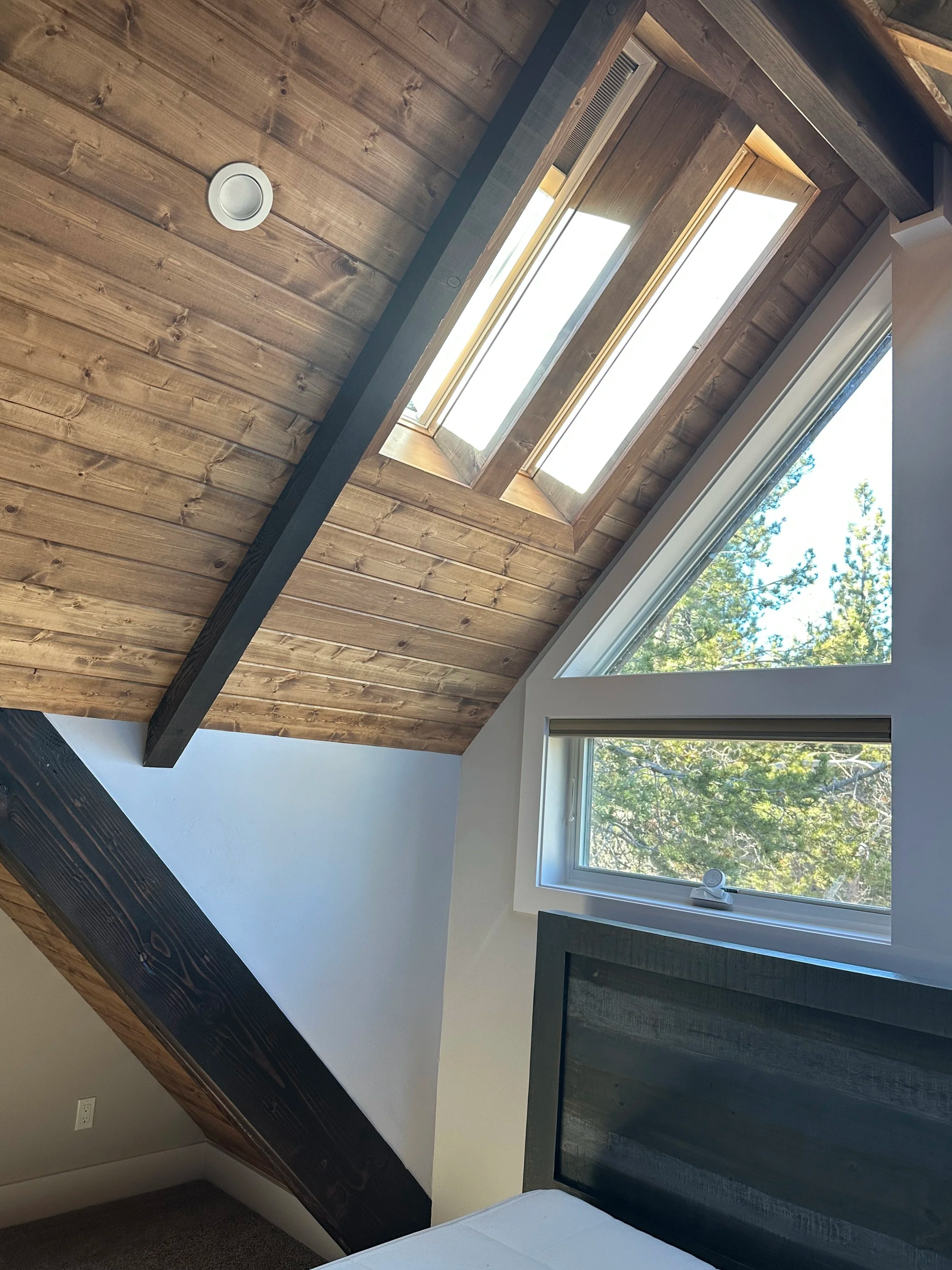 Interior view of a room with a sloped wooden ceiling featuring skylights, a window showing trees outside, and a dark wooden beam.