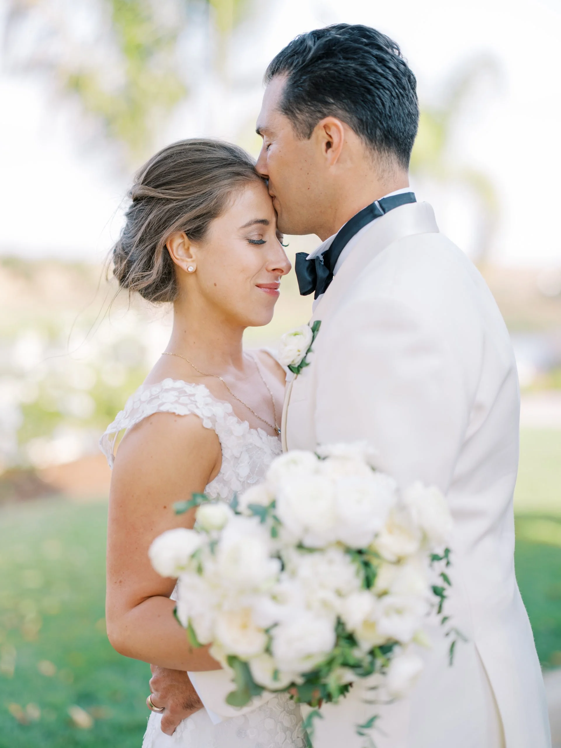 A newlywed couple; the groom is kissing the bride on her forehead while she holds a bouquet of white flowers and smiles.