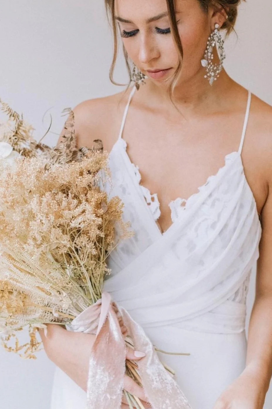 A woman in a white dress holding a bouquet of dried flowers, has long earrings, and is looking down.