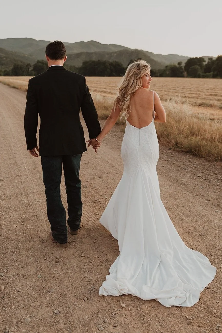 A bride and groom holding hands and walking down a dirt road in a rural area during sunset, with mountains in the background.