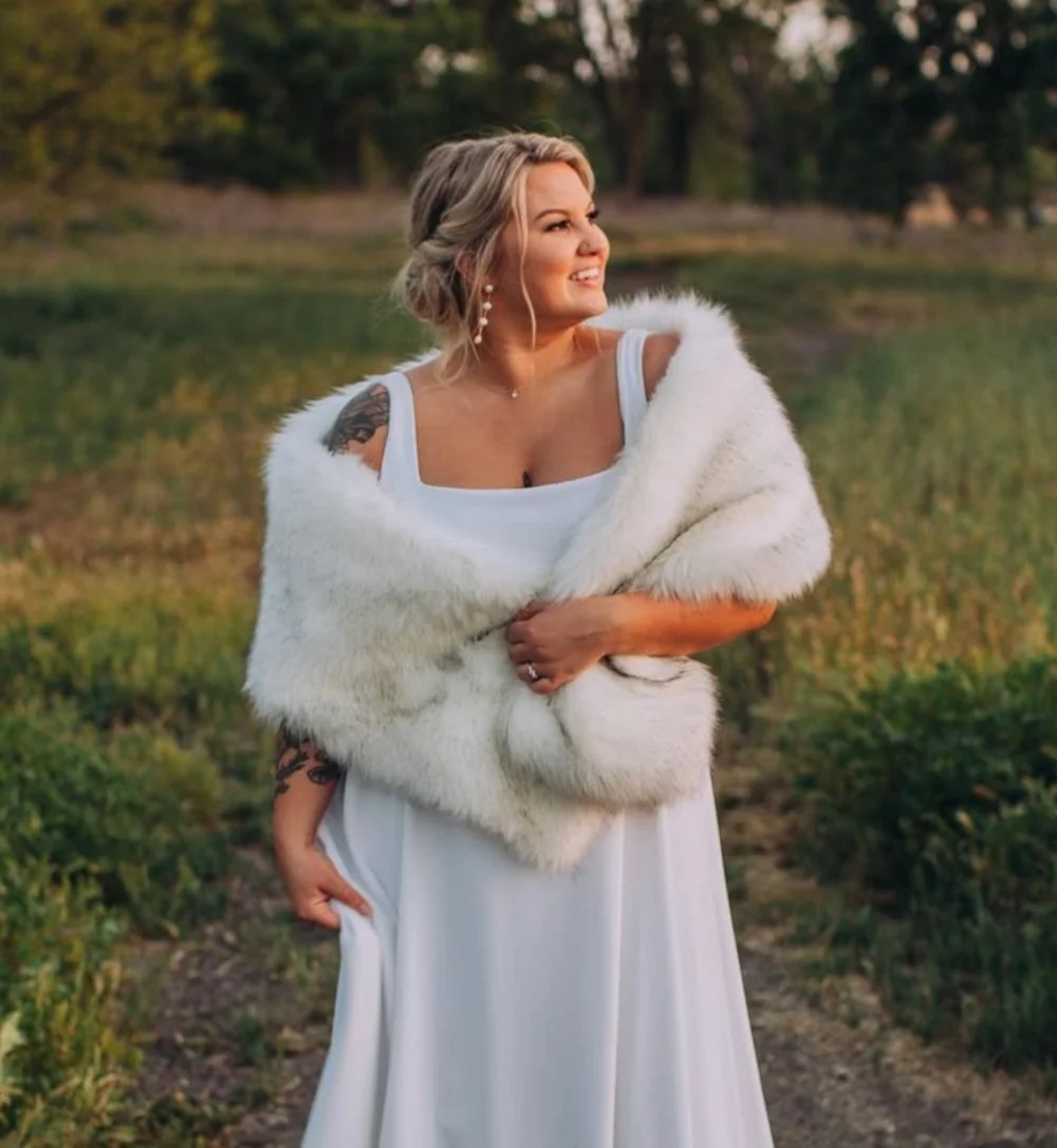 A woman in a white dress and fur shawl standing outdoors in a field with trees, smiling and looking to the side.
