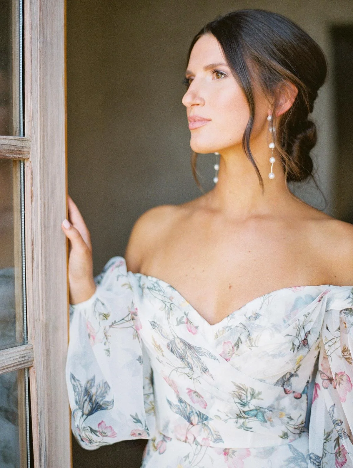 A woman with dark hair styled in an updo wearing pearl-drop earrings and an off-the-shoulder dress with a floral pattern, looking out a window.