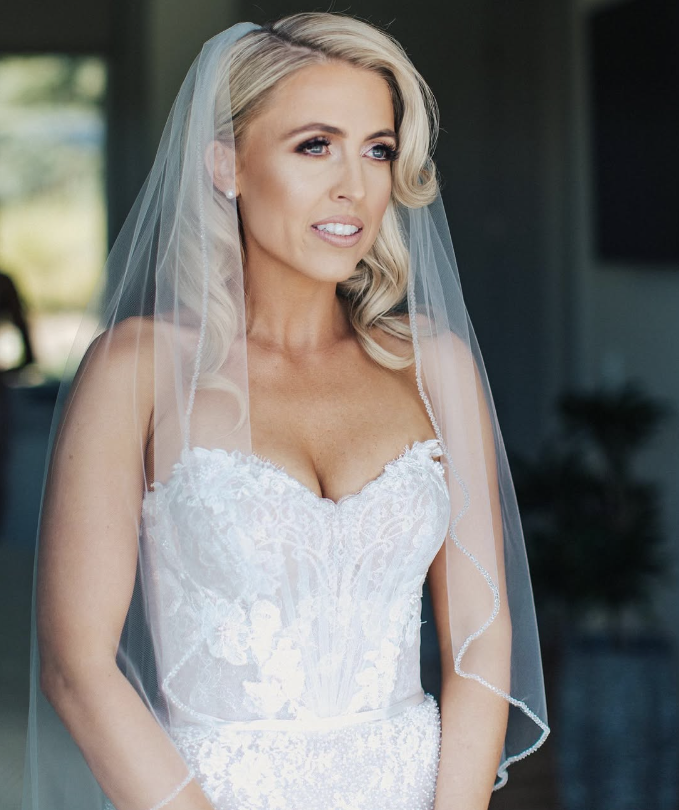 A bride in a white lace wedding dress with a sheer veil, standing indoors.