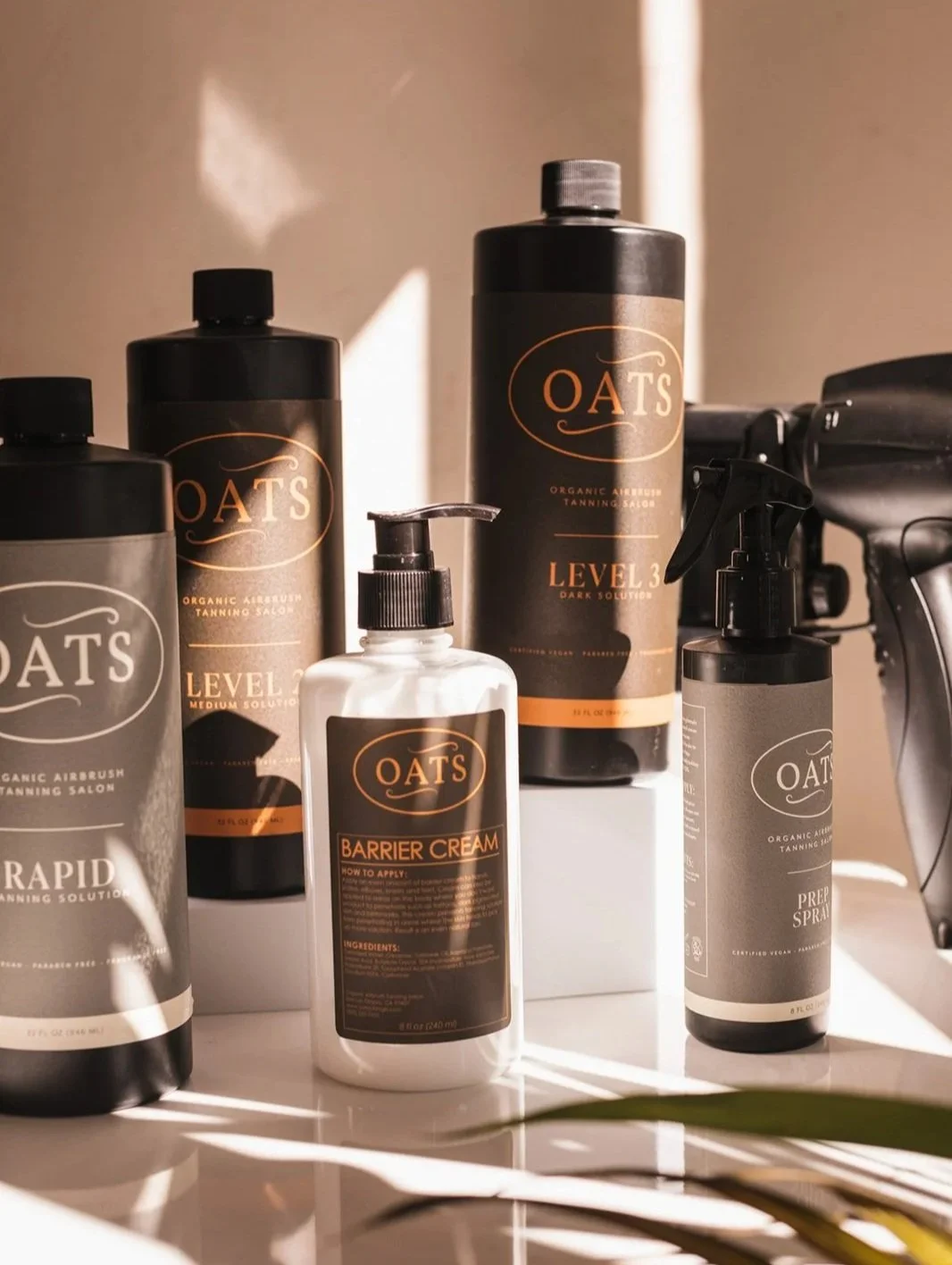 Various bottles of OATS organic airbrush tanning products, including a barrier cream, spray solutions, and a tanning lotion, on a white surface with sunlight shadows.