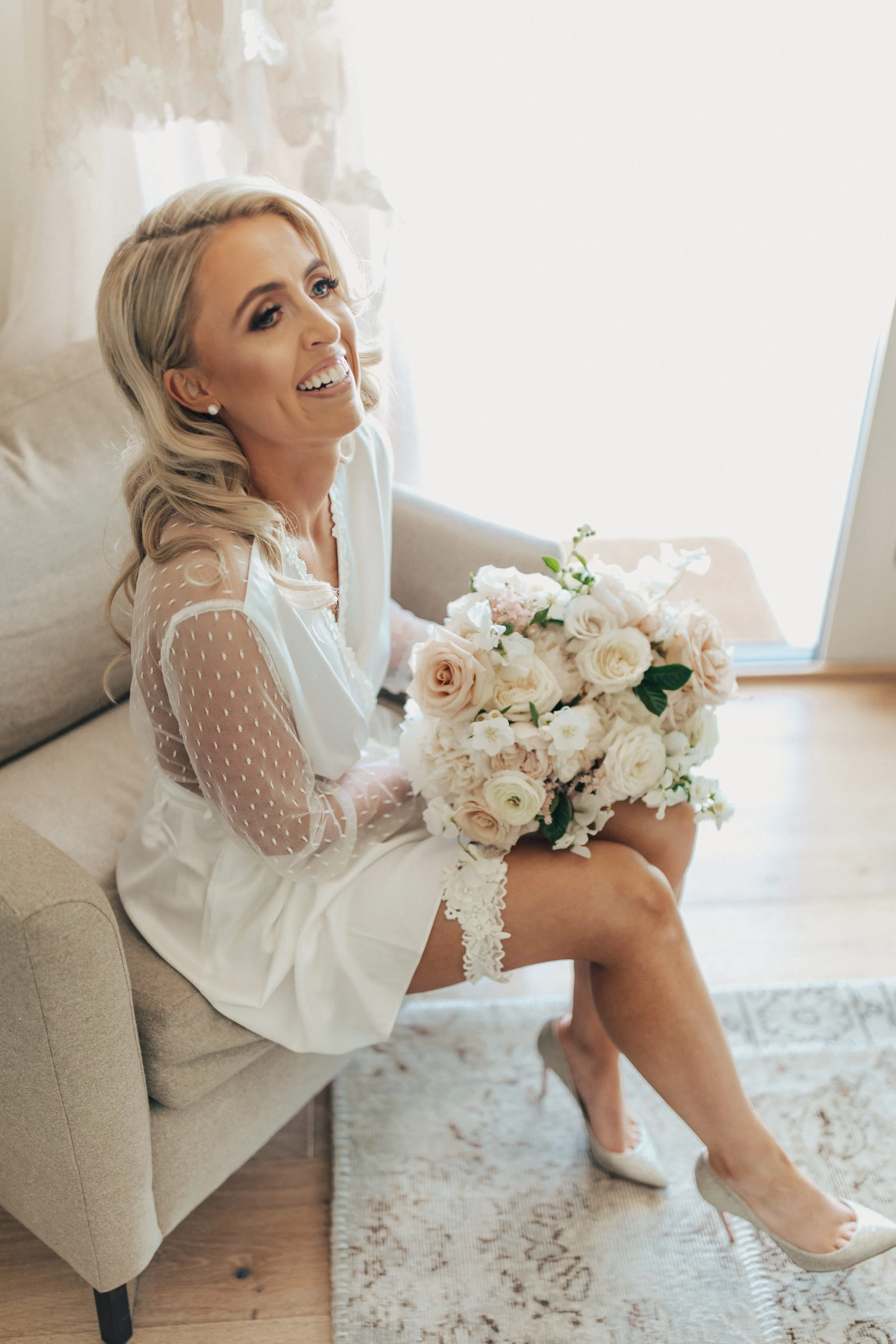 A smiling woman with blonde hair sitting on a beige sofa, holding a bouquet of white and blush roses, dressed in a white wedding robe with sheer polka dot sleeves, wearing white high heels, in a bright room with a window behind her.