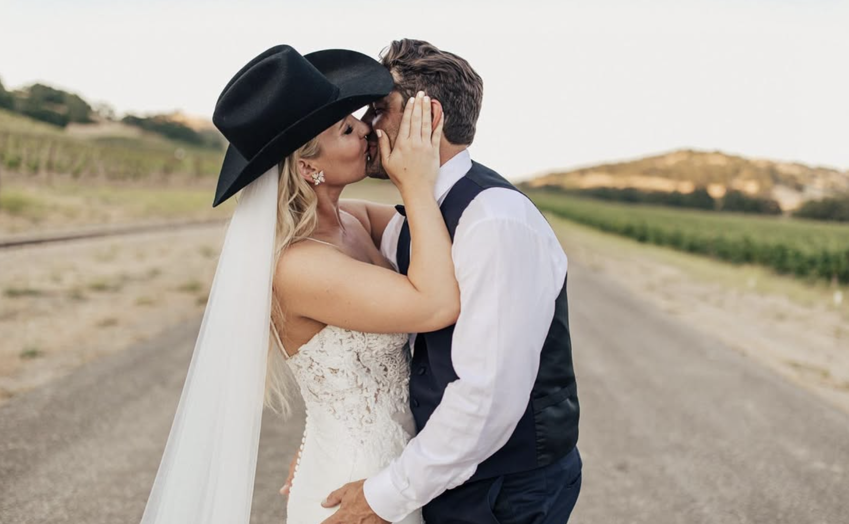 A bride and groom sharing a kiss outdoors, with the bride wearing a white lace dress, a black hat, and long veil, and the groom in a white shirt and black vest, in a rural setting with fields and hills in the background.
