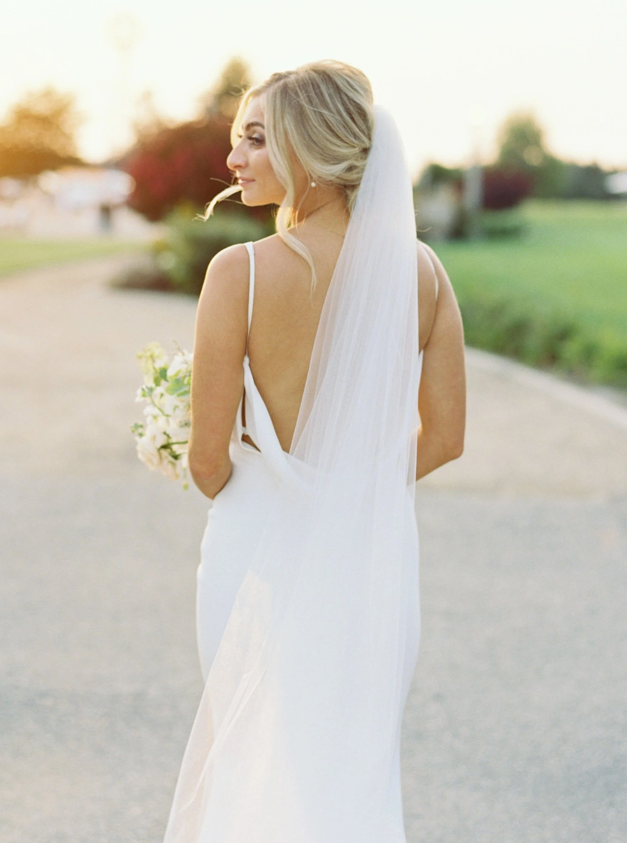 A woman in a white wedding dress and veil holding a bouquet of flowers, standing outdoors during sunset.