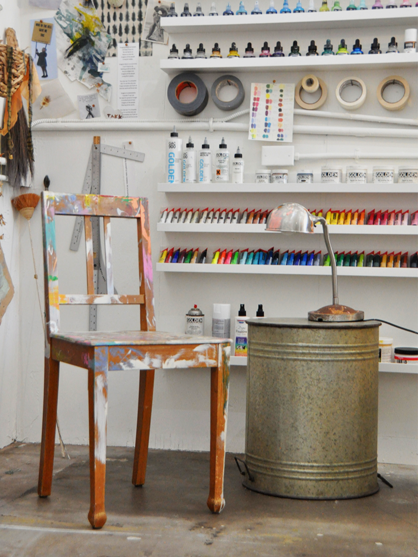 Art studio workspace with a paint-splattered chair, shelves of colorful paint and supplies, a desk lamp, and various art materials on shelves.