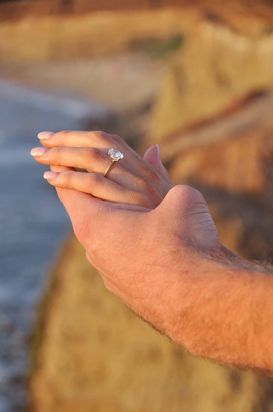 Close-up of a hand with a ring on the ring finger, held against a blurred natural background, possibly at sunset.