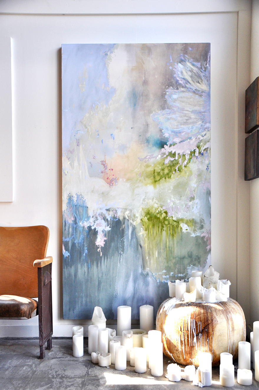 A large, abstract landscape painting with soft pastel colors, depicting a water scene with trees and sky, is mounted on a white wall. In front of the painting, there are white candles of various sizes placed on the floor and an unlit, round, wooden object, possibly a decorative piece or a torchiere, is also on the floor.