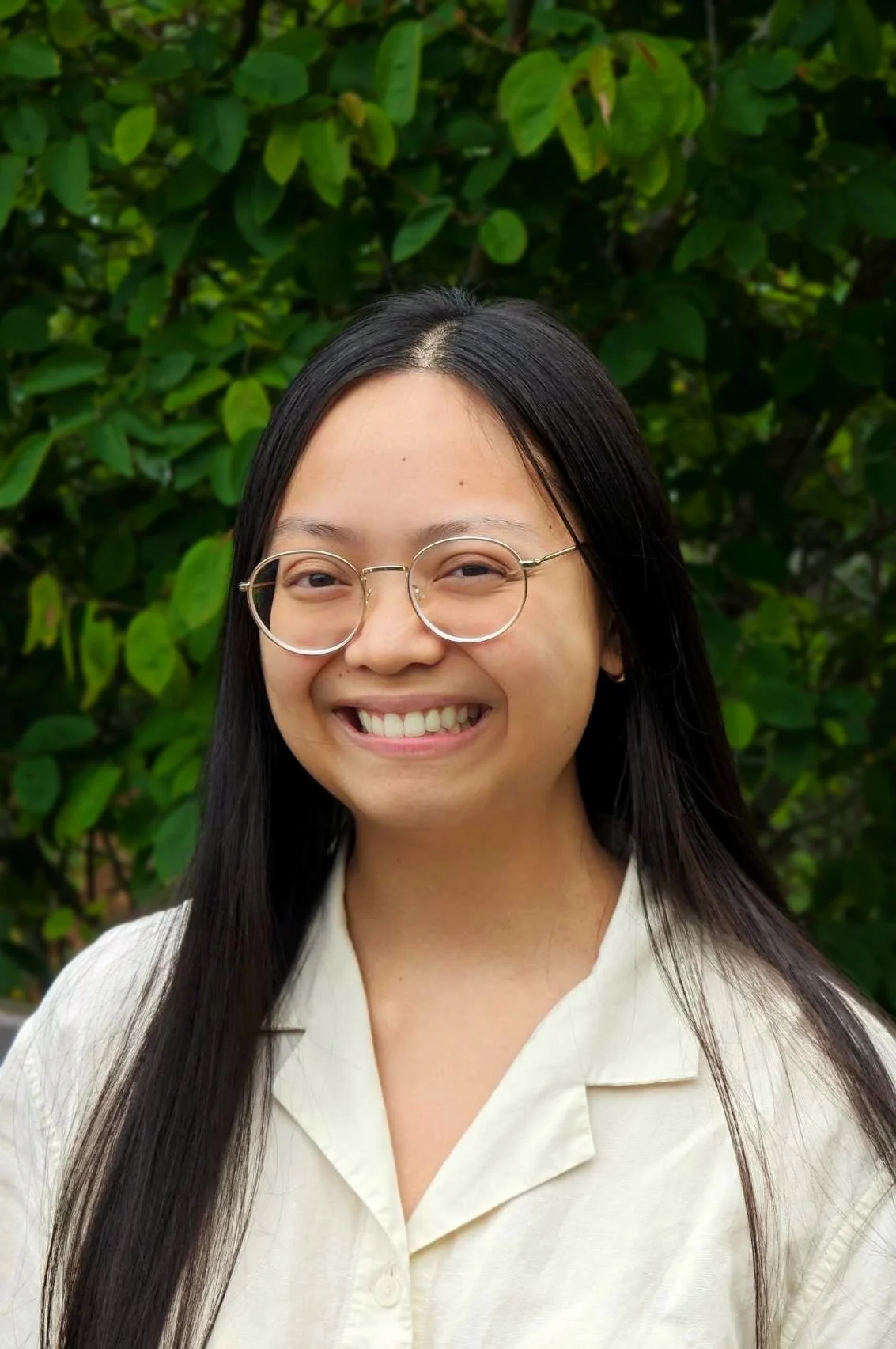 A woman with long black hair, wearing glasses and a white shirt, smiling outdoors in front of green leafy bushes.
