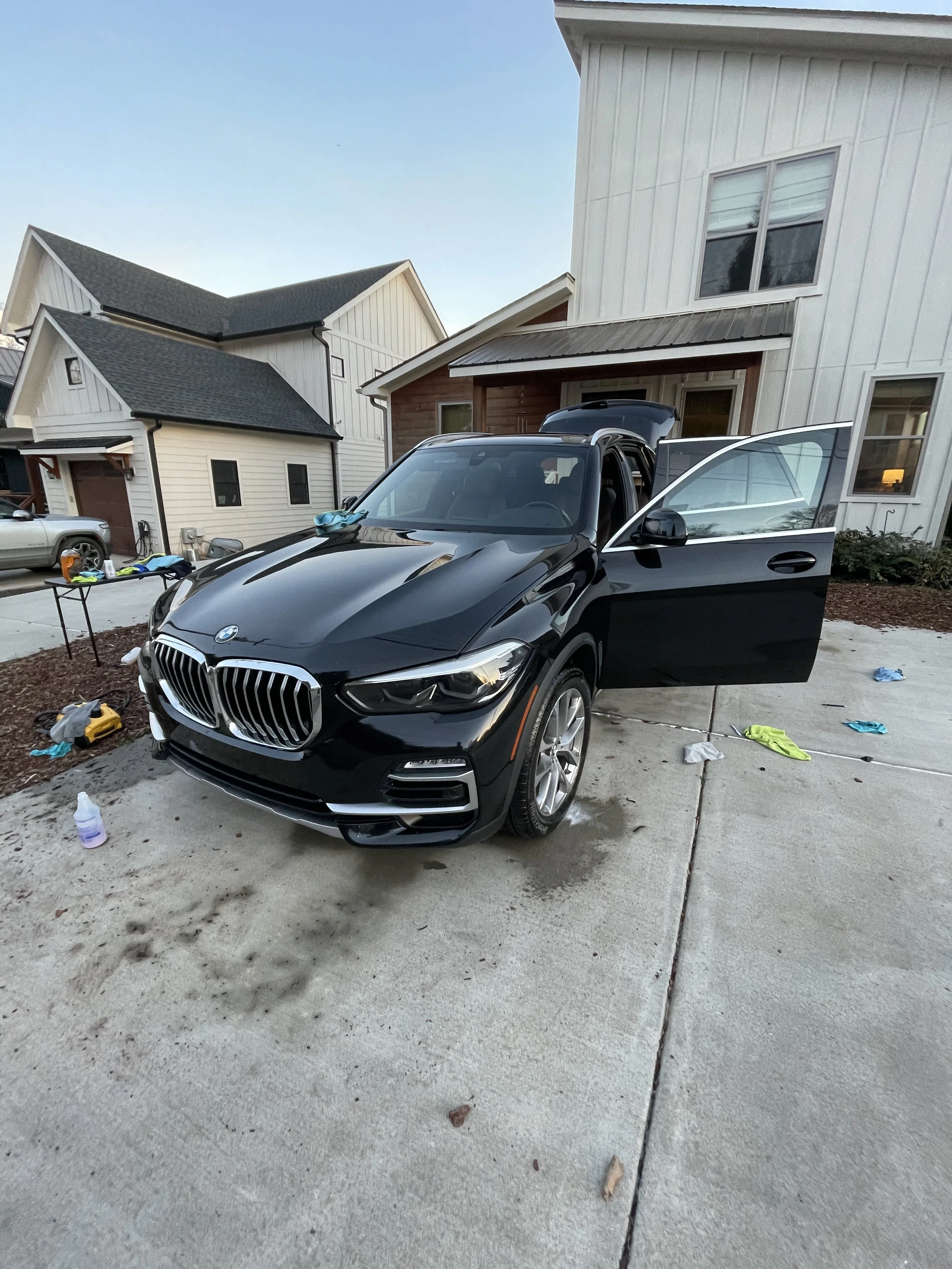 A black BMW SUV with an open passenger door parked in a driveway outside a white modern house, with various cleaning supplies and clothes scattered on the ground.