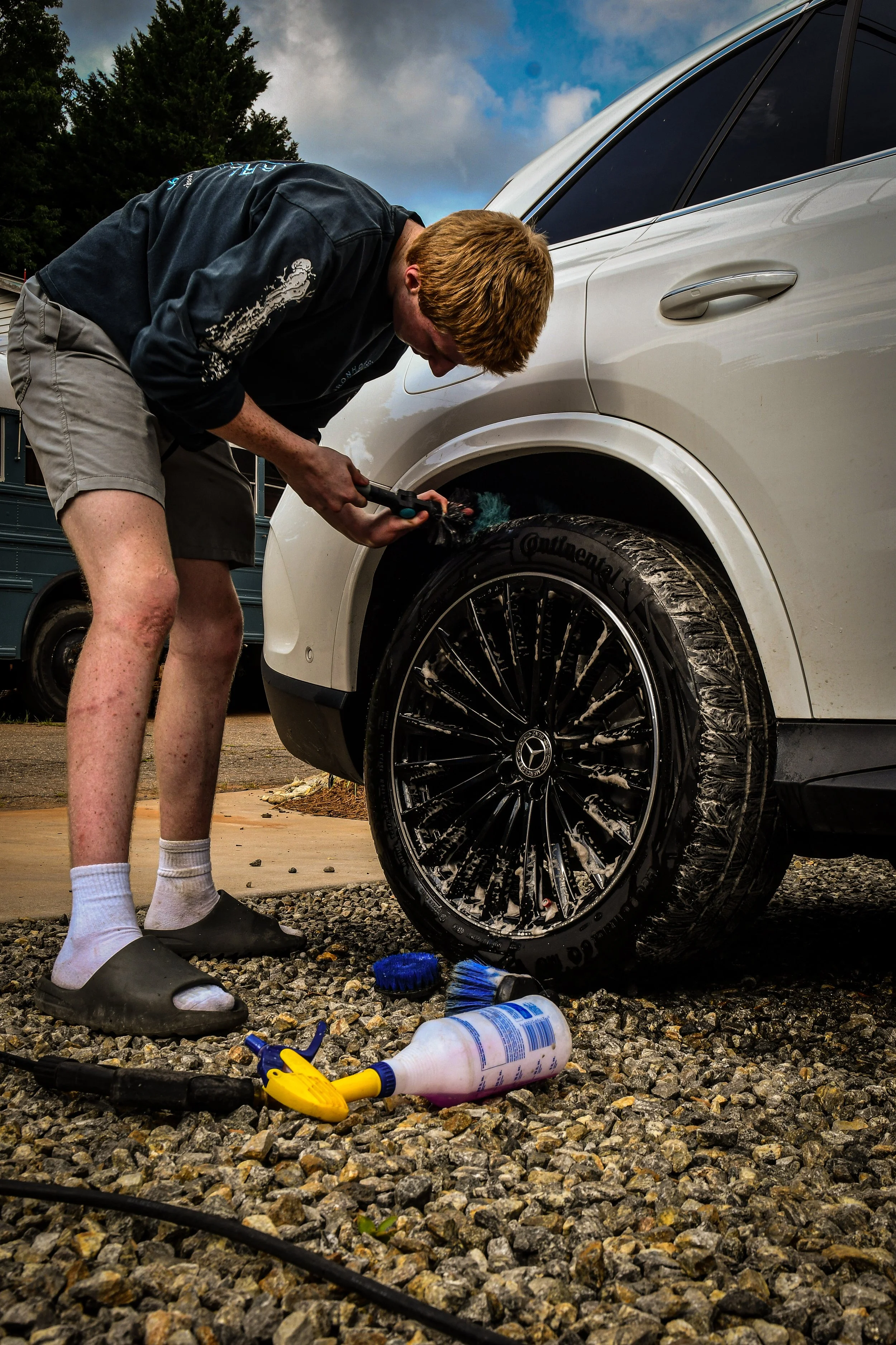 A man washing his car's front tire with a power washer, standing on a gravel driveway with car cleaning supplies nearby.