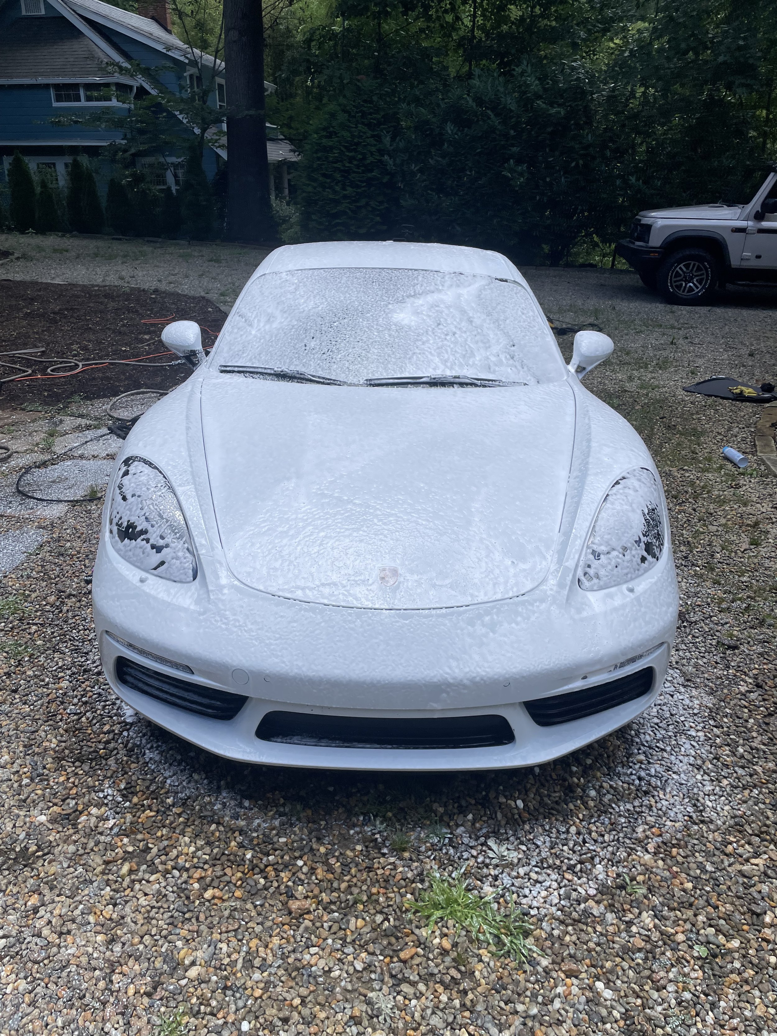 Front of a white sports car covered in foam, being washed outdoors on a gravel driveway.