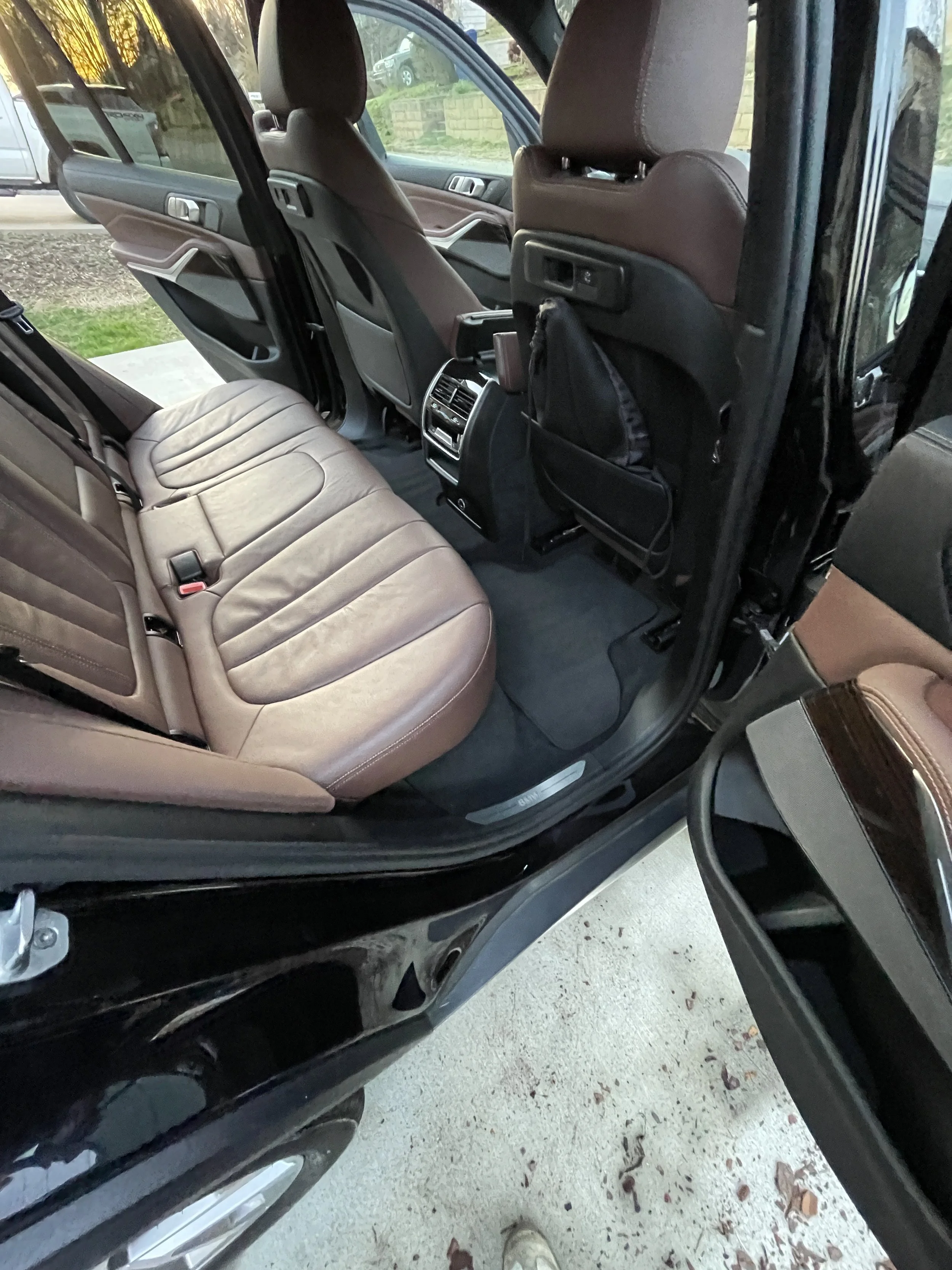 The interior of a black SUV, showing brown leather seats, the back of the front seats with seatback pockets, and the rear bench seat. The car door is open, revealing a driveway with leaves on the ground.