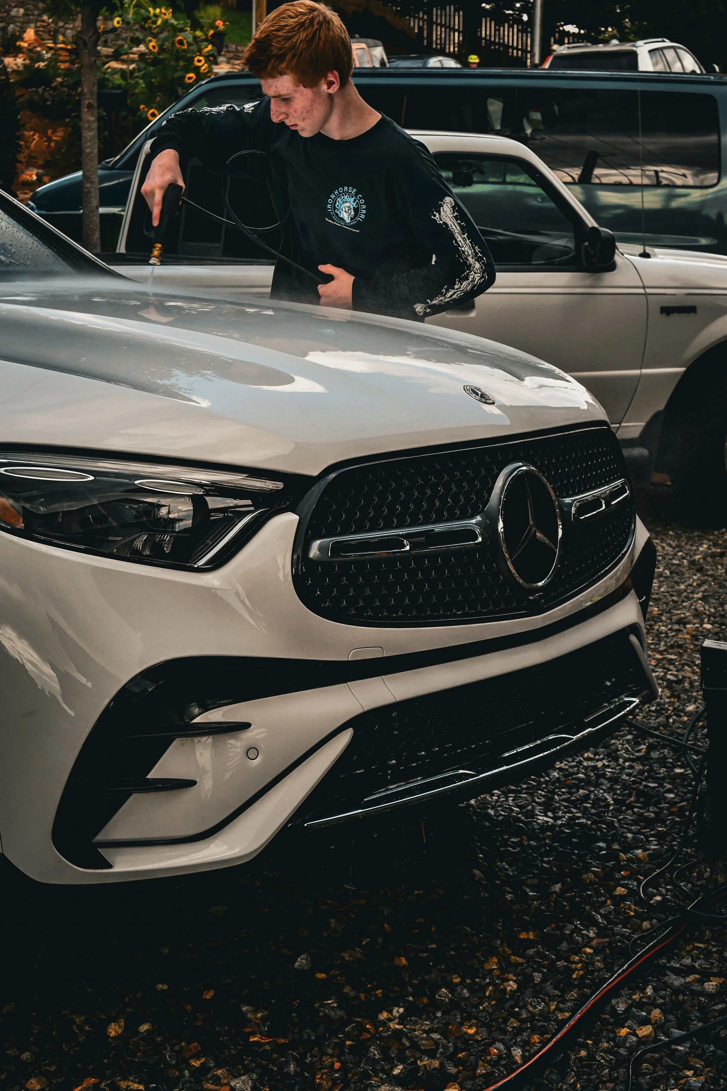 A young man with red hair and freckles is washing a white Mercedes-Benz car in an outdoor parking lot. He is wearing a black long-sleeve shirt and holding a hose or spray gun, cleaning the vehicle. Several other parked cars are visible in the background.