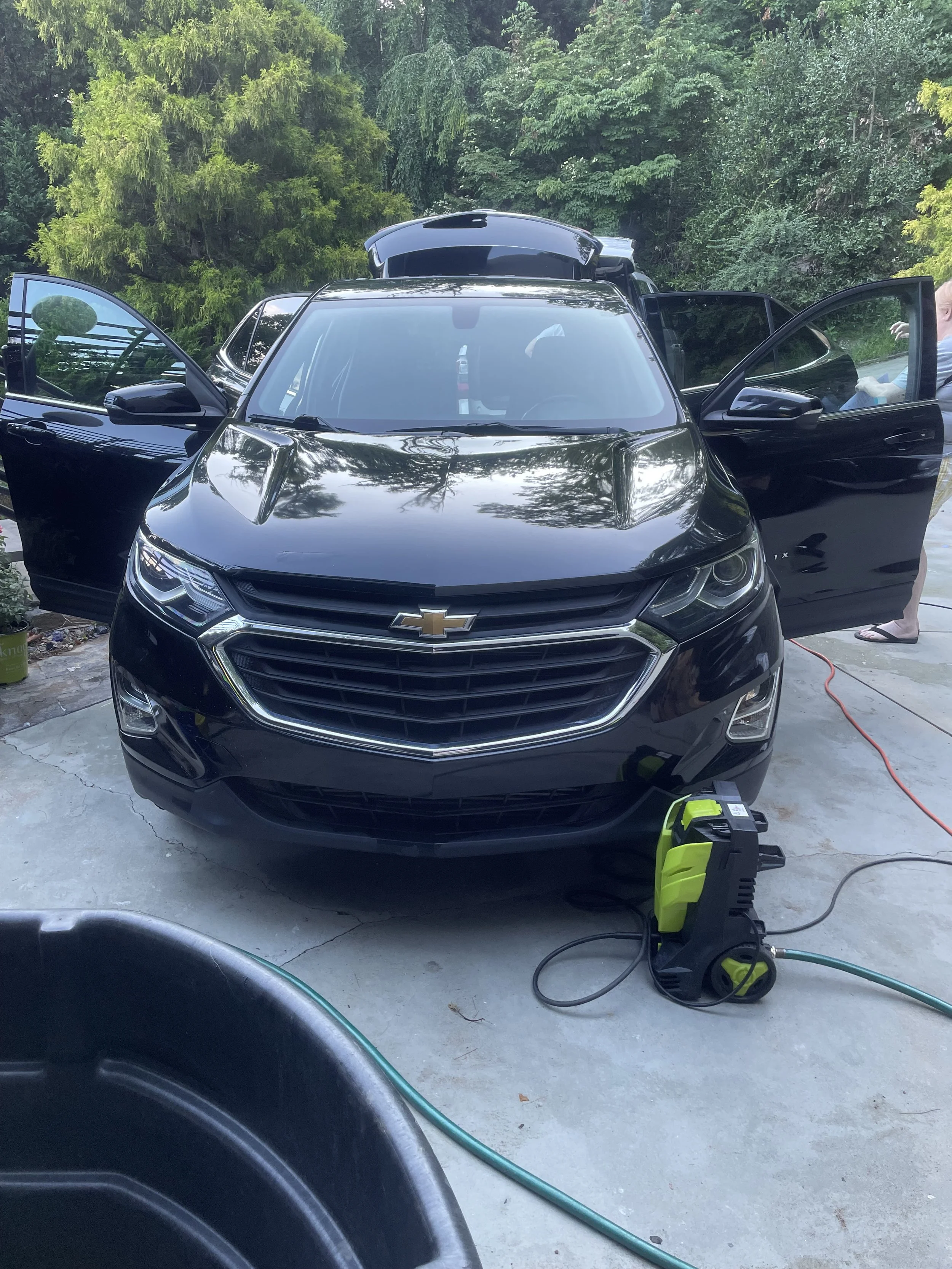 Front view of a black Chevrolet SUV with the doors open, parked outdoors on a concrete driveway. A person is visible on the right side, and a green pressure washer is placed on the ground in front of the vehicle. There are trees and greenery in the background.