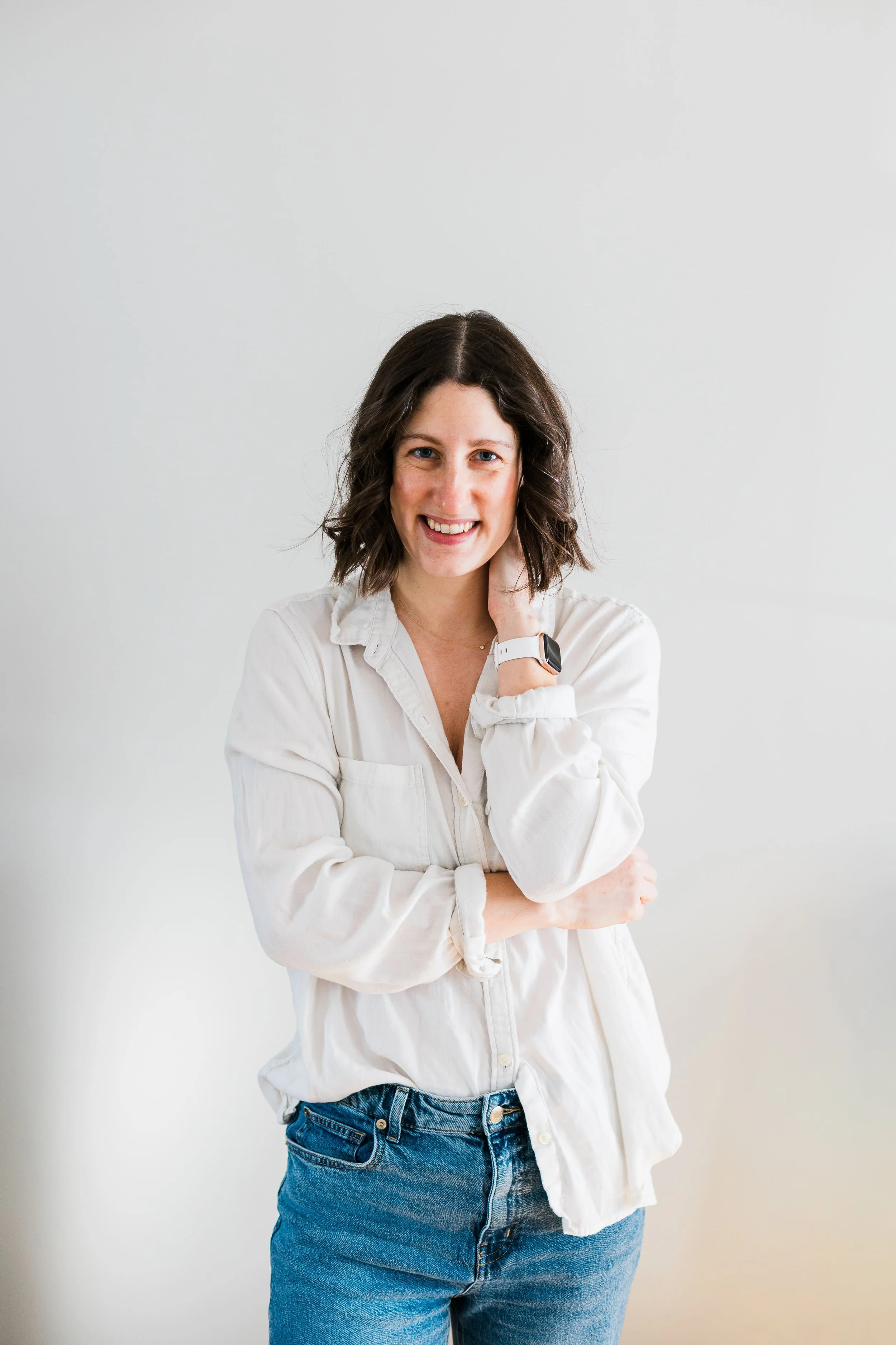 Woman with dark brown hair, white shirt and blue jeans against a light grey wall, smiling with one hand around her neck