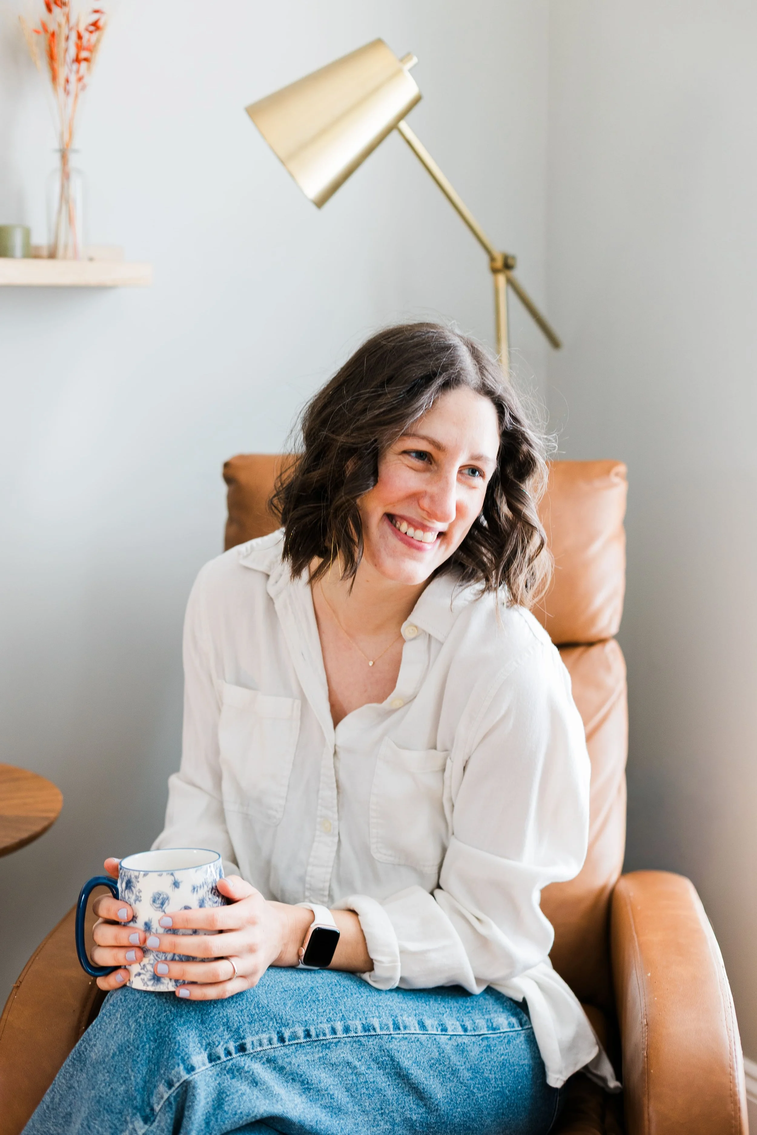 woman with dark brown hair, smiling, wearing a white button down shirt and jeans, sitting in a brown chair and holding a cup