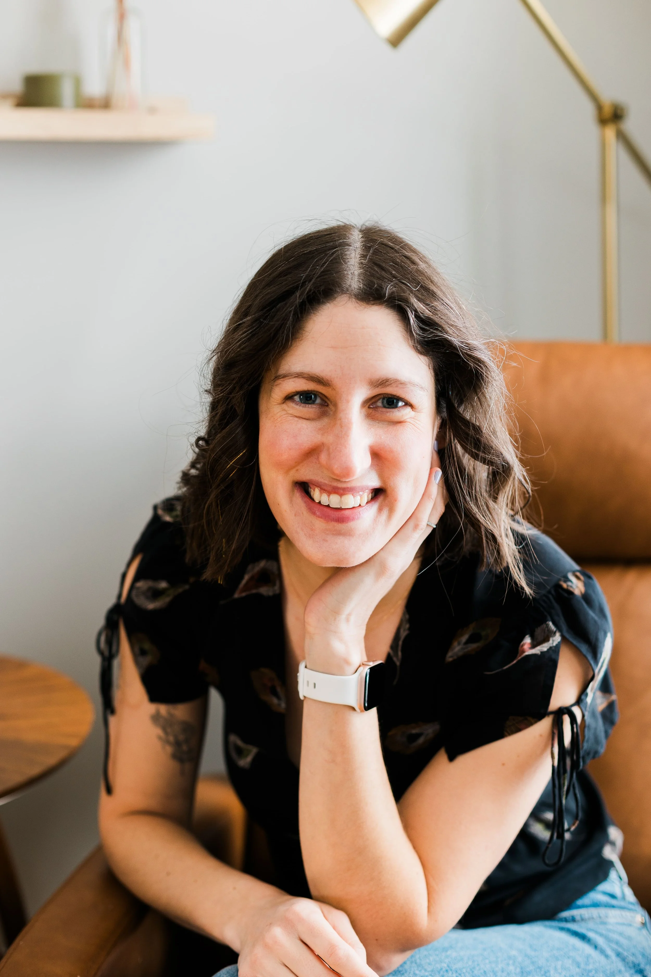 woman with dark brown hair, wearing a black shirt, posing in a brown chair, with a smile